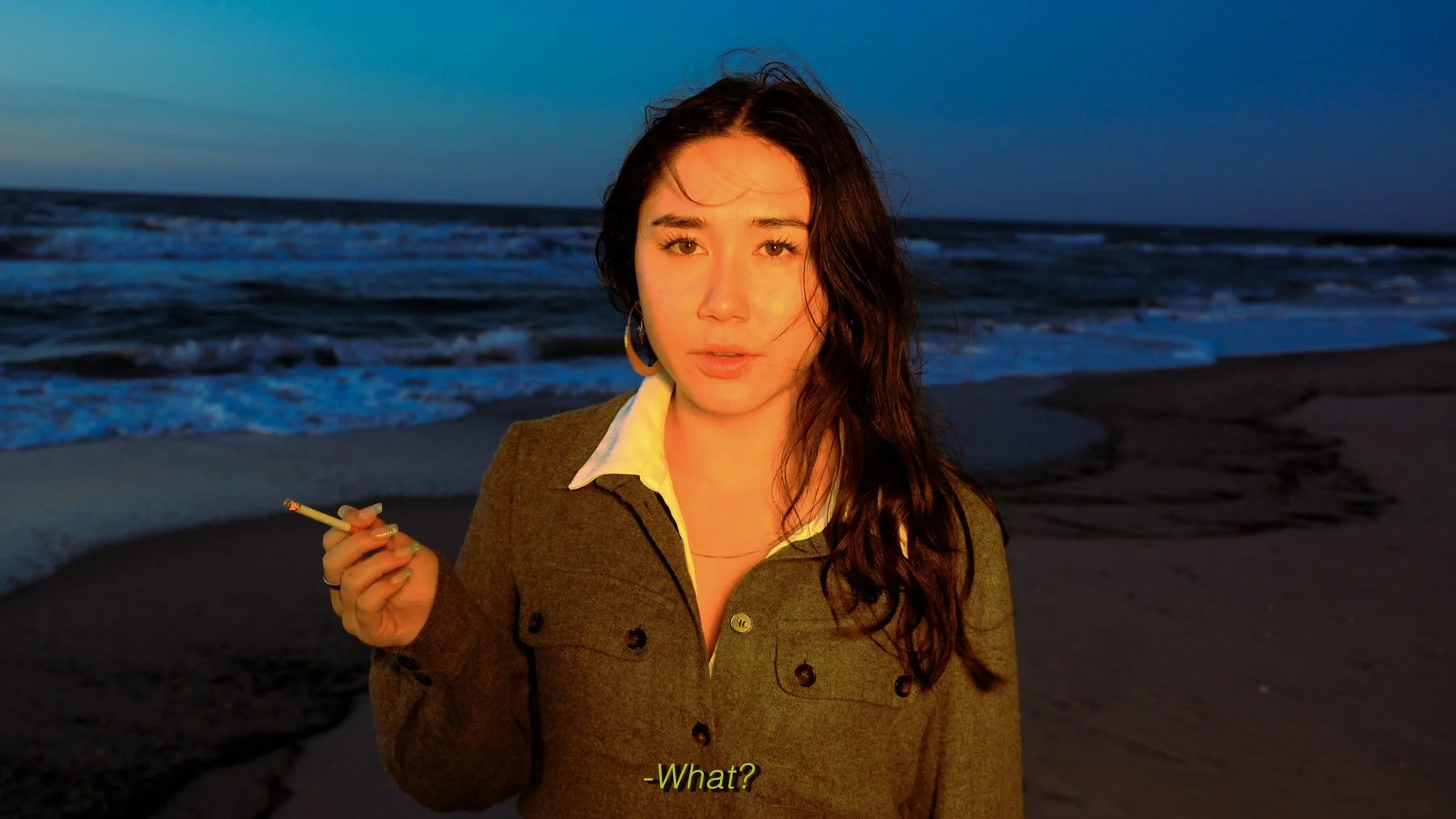 A woman with dark hair, wearing a brown jacket and white shirt, stands on a beach during sunset or dusk, holding a cigarette and speaking with the subtitle '-What?' at the bottom.