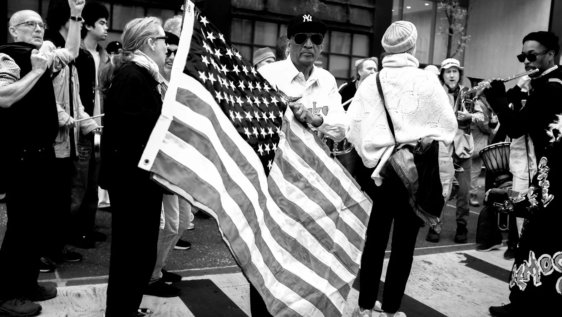 A group of people participating in a protest or demonstration, with one man holding a large American flag and others around him, some playing musical instruments.