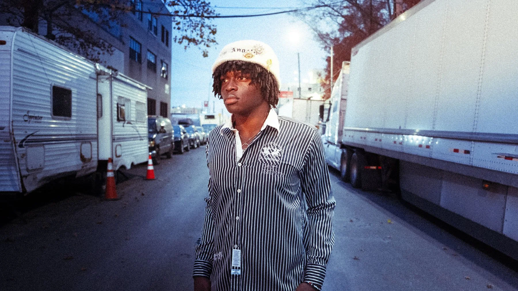 A young man with curly hair wearing a black and white pinstripe shirt and a white beanie stands on a city street at dusk, with parked cars and trucks behind him.