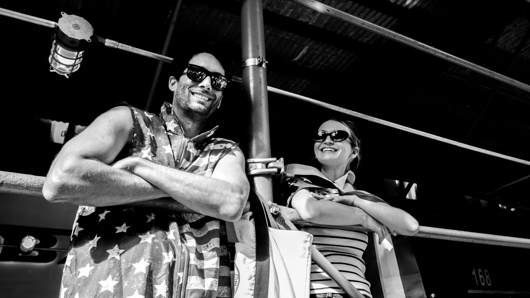 Two people, a man and a woman, are leaning on a railing and smiling. They are wearing sunglasses, and the man is dressed in a sleeveless jacket with an American flag pattern. The woman is wearing a striped shirt. There are pipes and a ceiling in the 