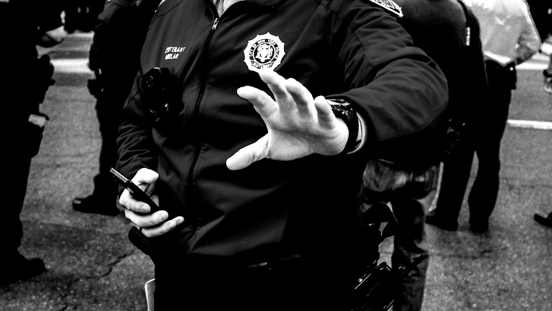 Close-up of police officer with badge, holding a phone in one hand and raising the other hand in a stop gesture during a protest or crowd control event.