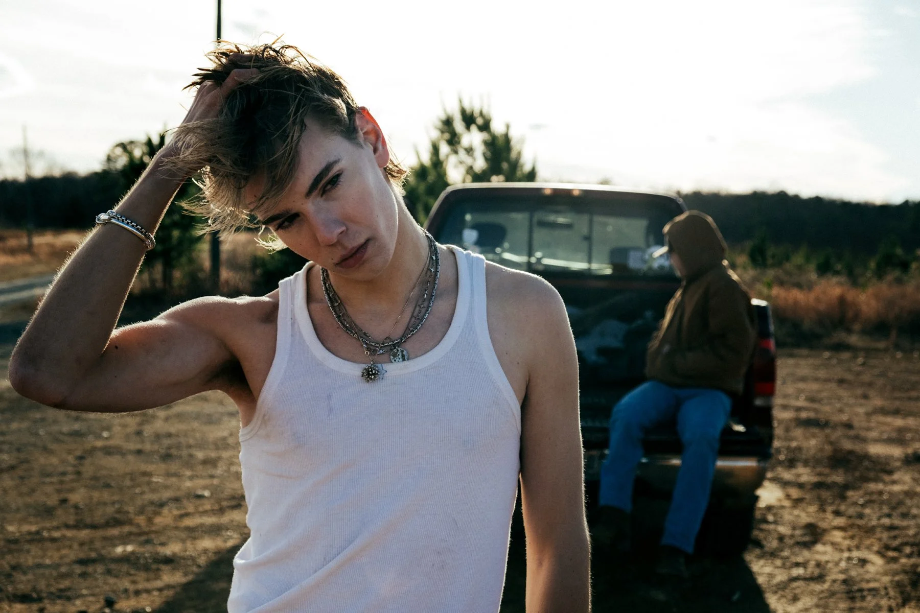 A young woman with short, messy hair, wearing a white tank top and layered necklaces, stands in the foreground with her hand running through her hair, looking directly at the camera. In the background, a person sits in the open tailgate of a pickup t