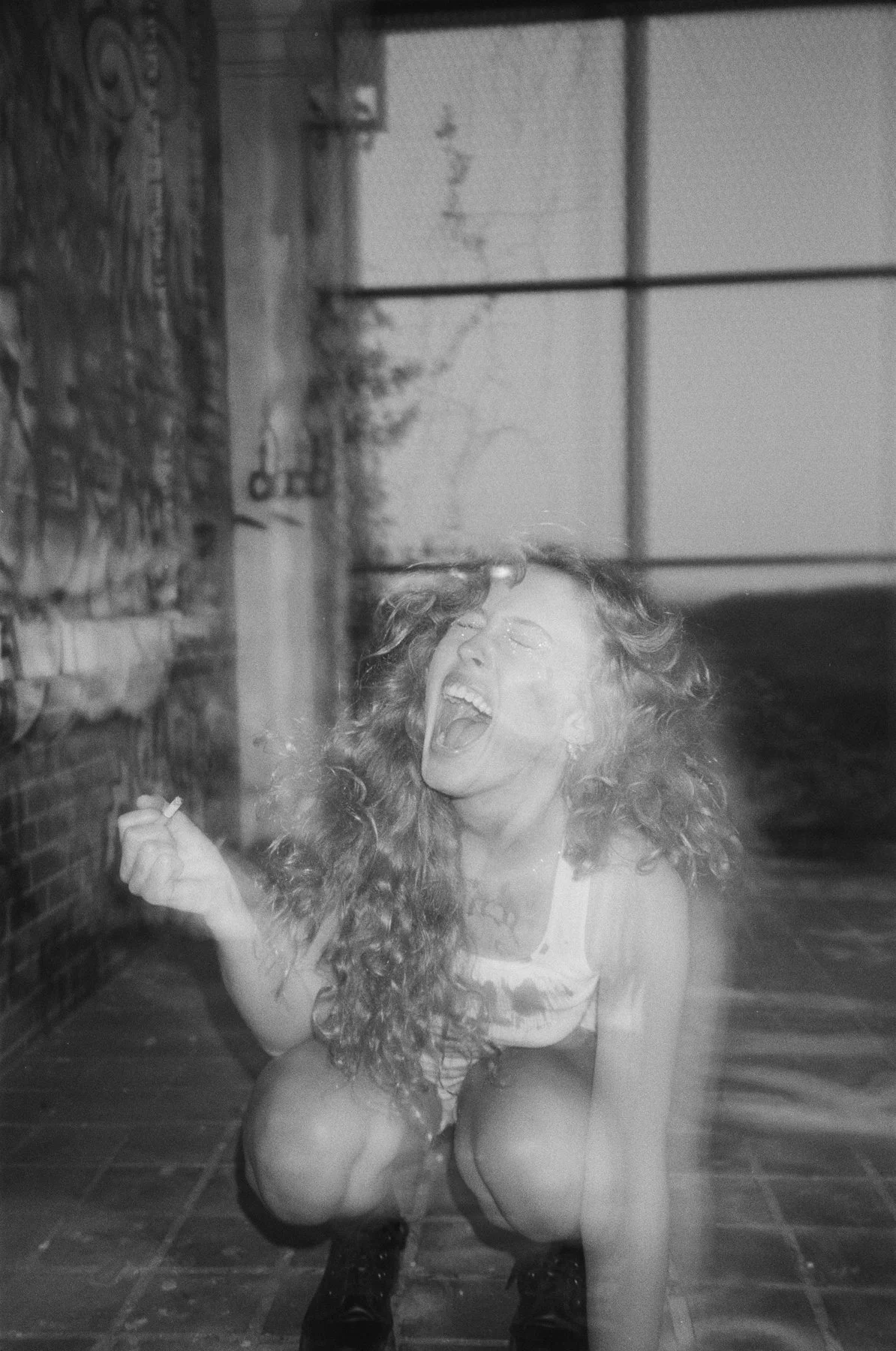 A woman with curly hair squatting on a tiled floor, smiling and laughing with her mouth open, holding a cigarette in her right hand. She appears to be in an industrial or urban setting.
