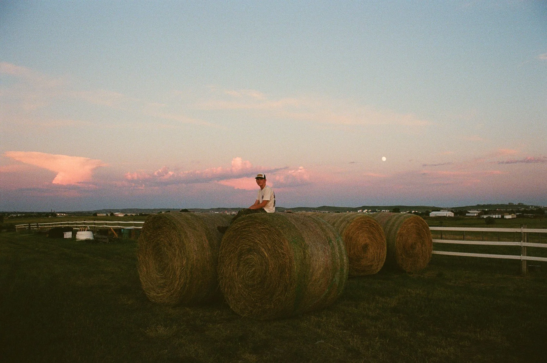 A young man sitting on large hay bales in a grassy field during sunset with a pastel-colored sky and full moon in the background.