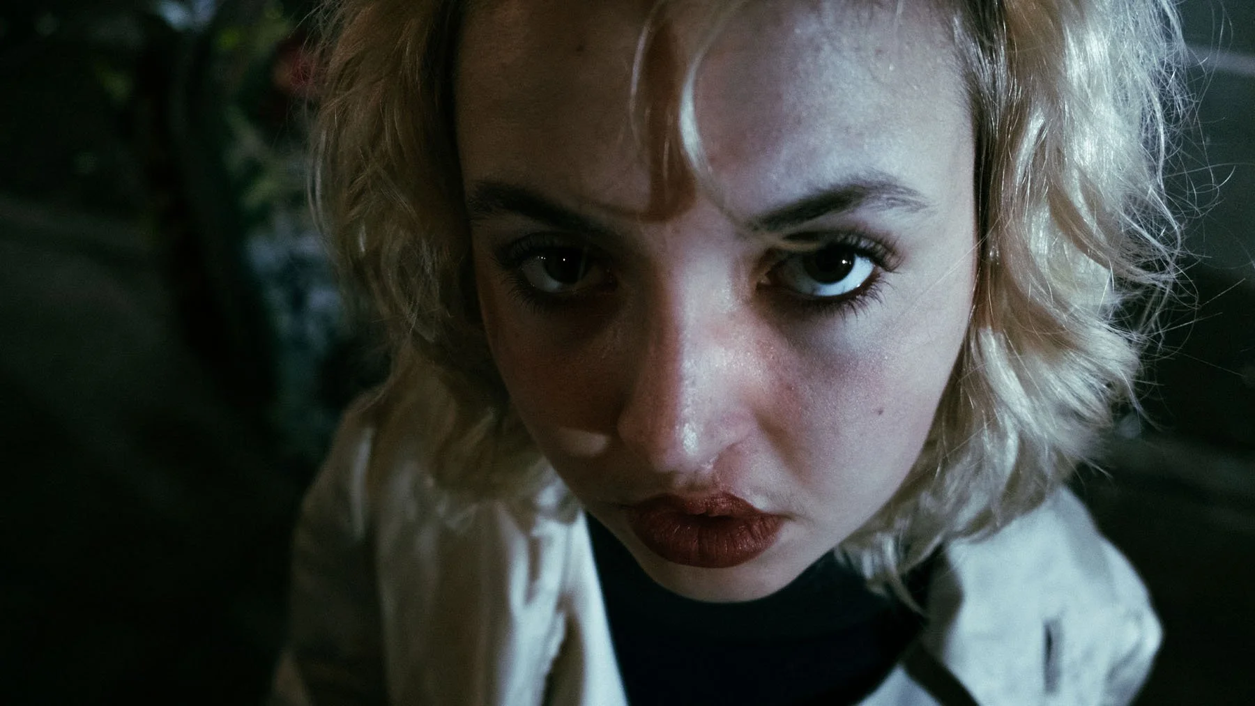 A young woman with curly blond hair and dark makeup looking directly into the camera, with a serious expression.