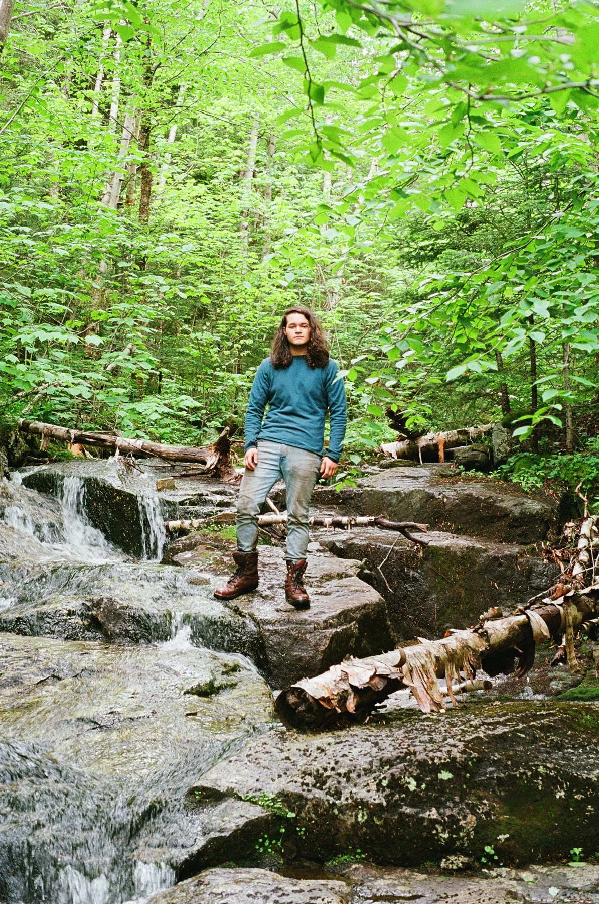 A person with long curly hair stands on a large rock in a forest next to a small waterfall, surrounded by green trees and foliage.