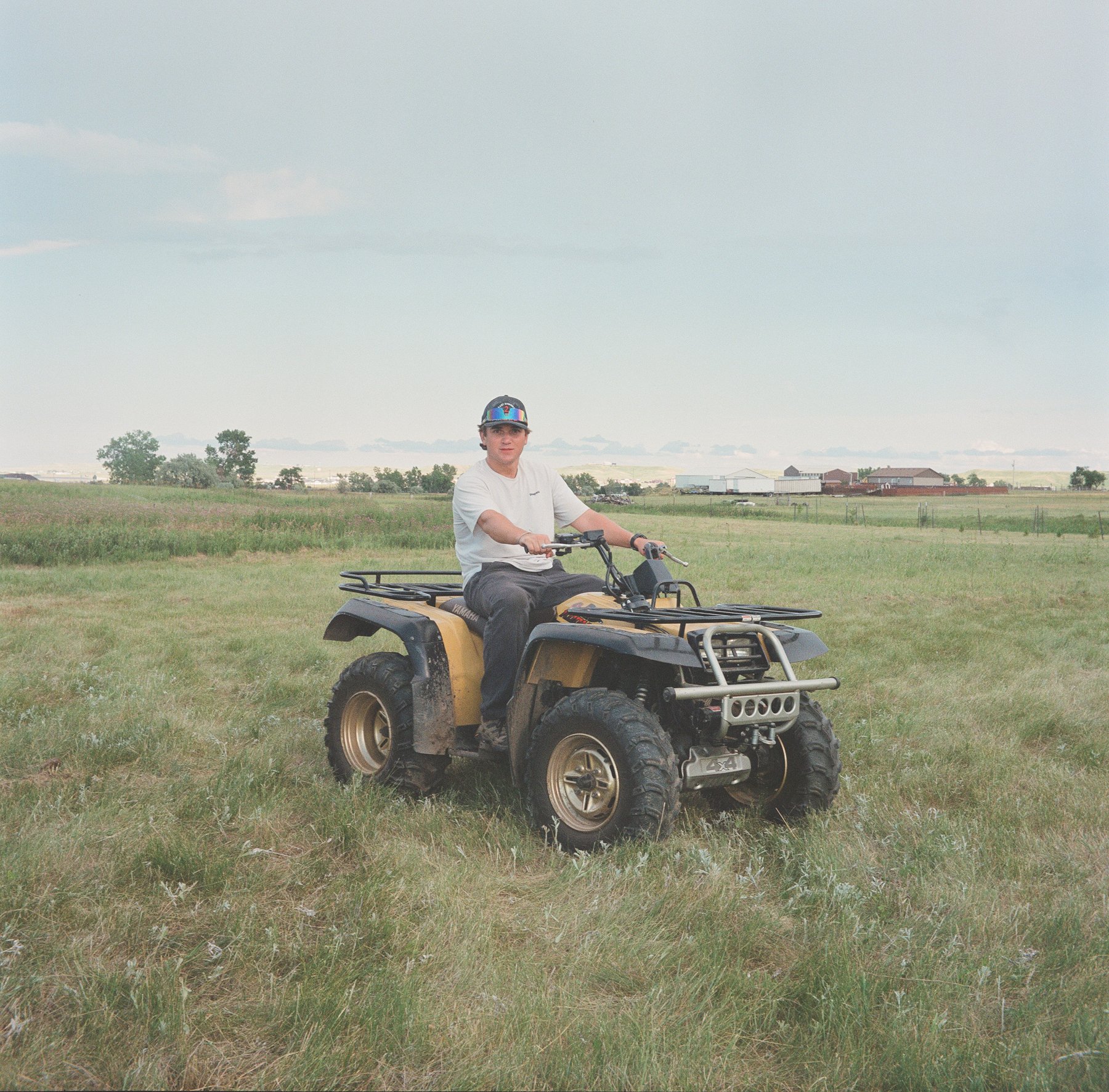 A young man wearing a helmet and white t-shirt riding a yellow and black all-terrain vehicle in an open grassy field with farm buildings in the background under a cloudy sky.