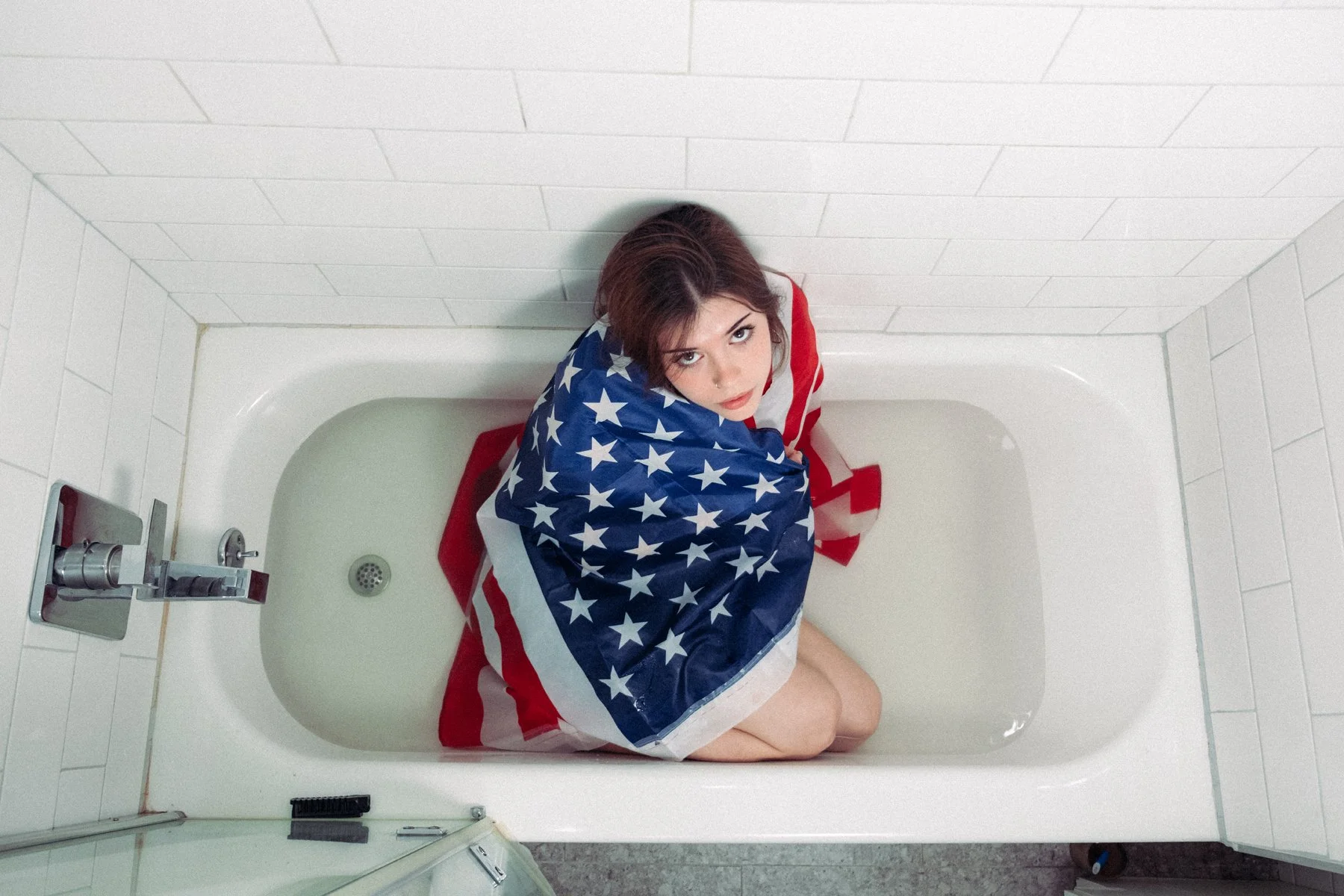 A woman sitting on a bathtub wearing an American flag as a blanket, looking up at the camera.