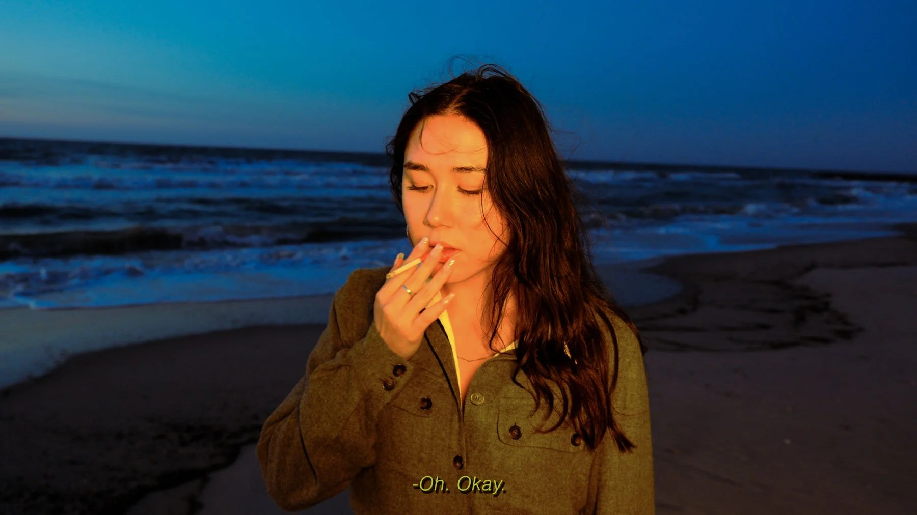 A woman with long, wavy hair stands on a beach during sunset, holding a cigarette near her lips with her eyes closed, and the ocean waves and sky in the background.