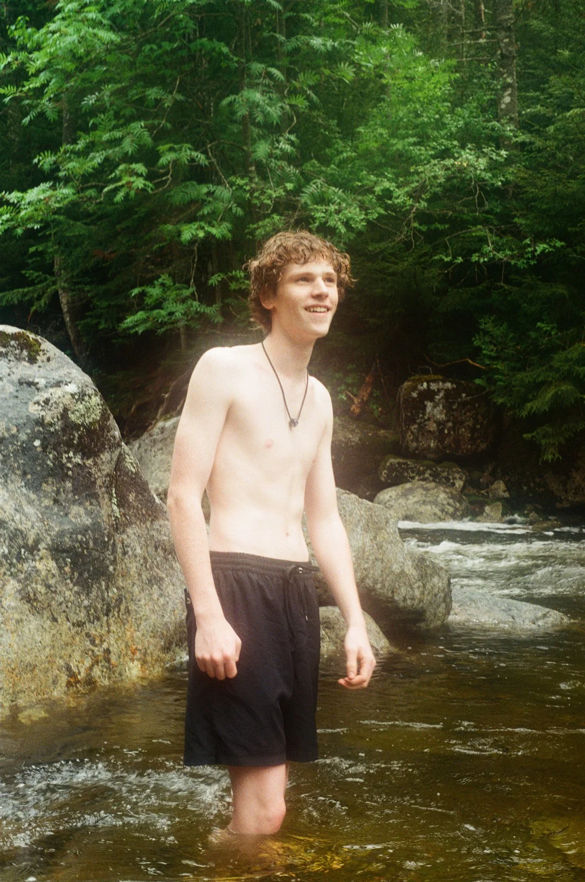A young man with curly hair is standing in a river, smiling, surrounded by large rocks and dense green trees.