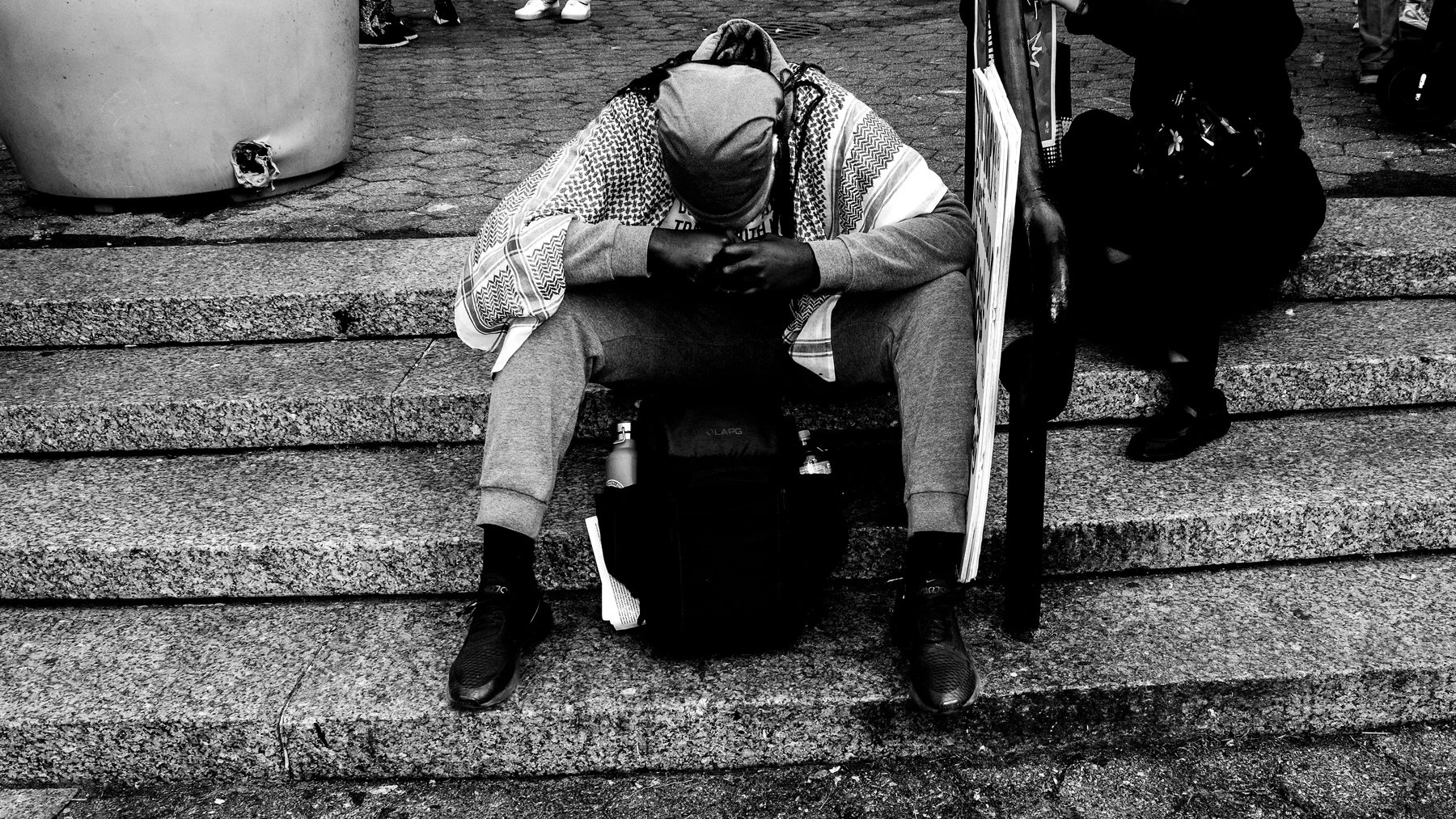 A man sitting on stairs, wearing a hooded jacket and sweatpants, looking at his phone, with a backpack beside him. A woman in black clothing is walking past on the steps.