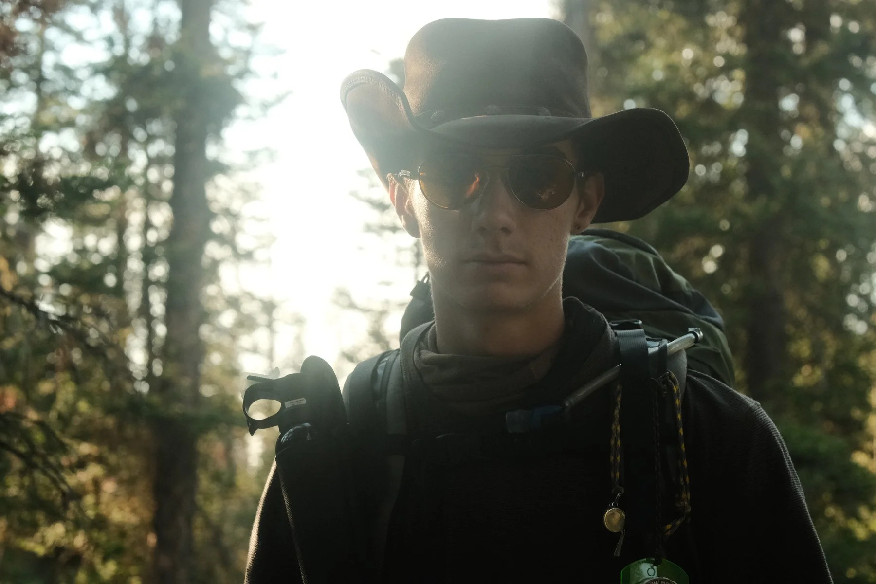 A person wearing a wide-brimmed hat, sunglasses, and outdoor gear with a backpack, standing in a forest with sunlight shining through trees behind them.
