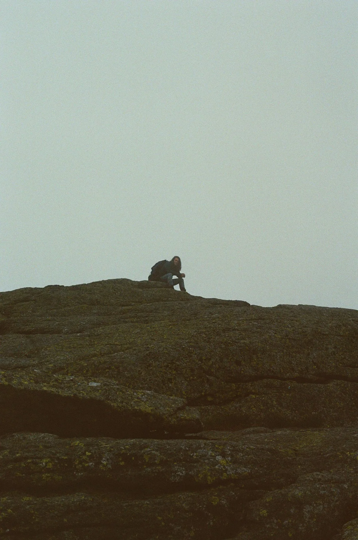 Person sitting on a large rocky surface outdoors, wearing a backpack, against a cloudy sky.