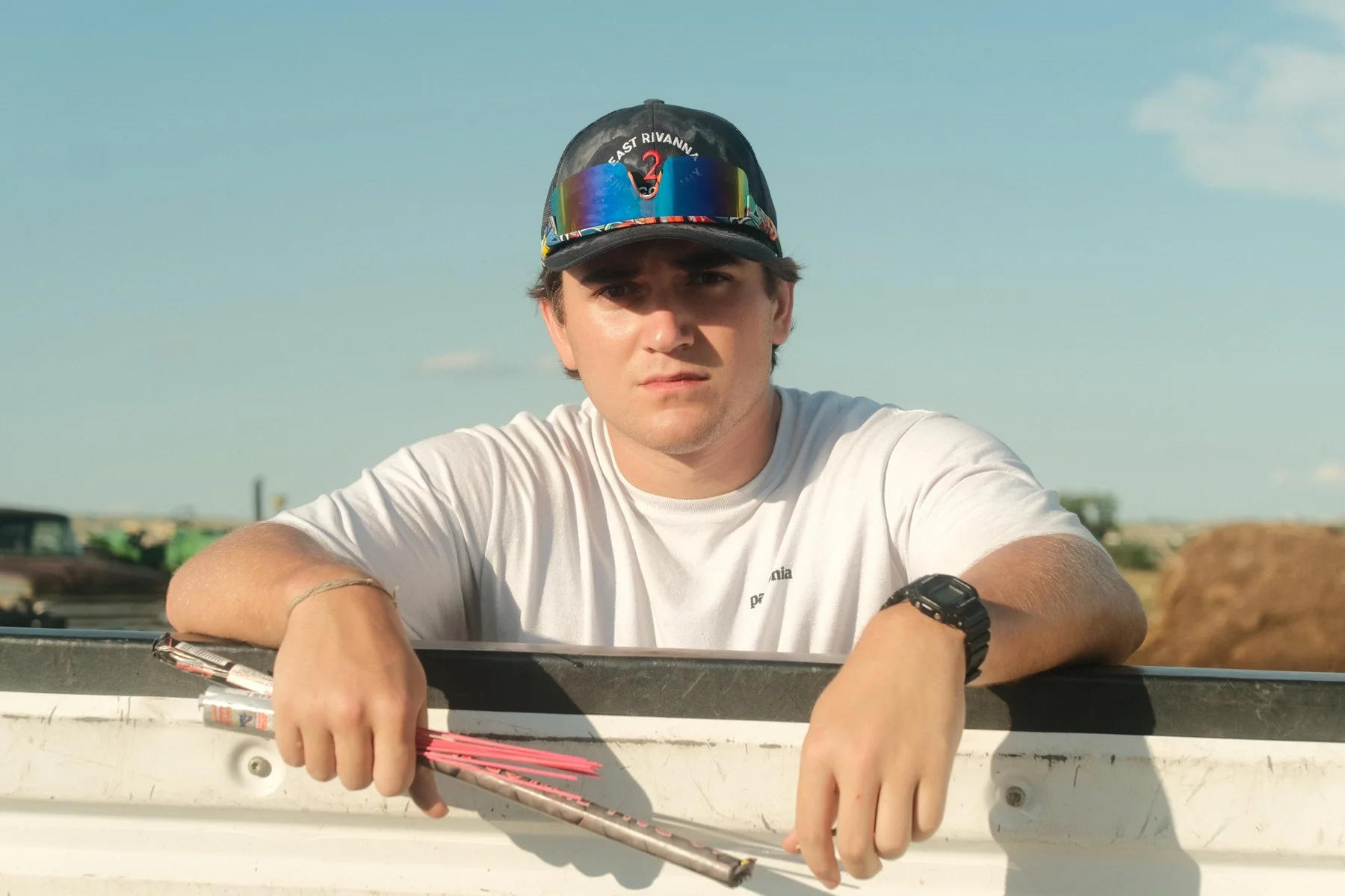 A young man wearing a black cap with a rainbow-colored reflective visor and a white T-shirt leaning on a boat, holding arrows, with a blue sky and some blurred outdoor objects in the background.