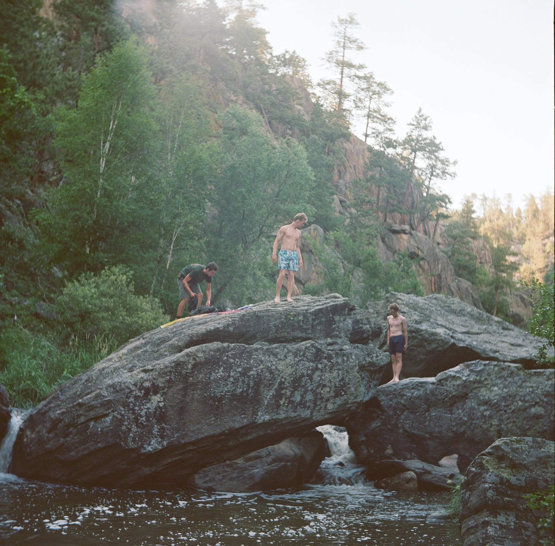 Three young men at a rocky waterfall in a forest, two shirtless and one dressed in a t-shirt, with two of them standing on rocks and one crouching near a backpack.