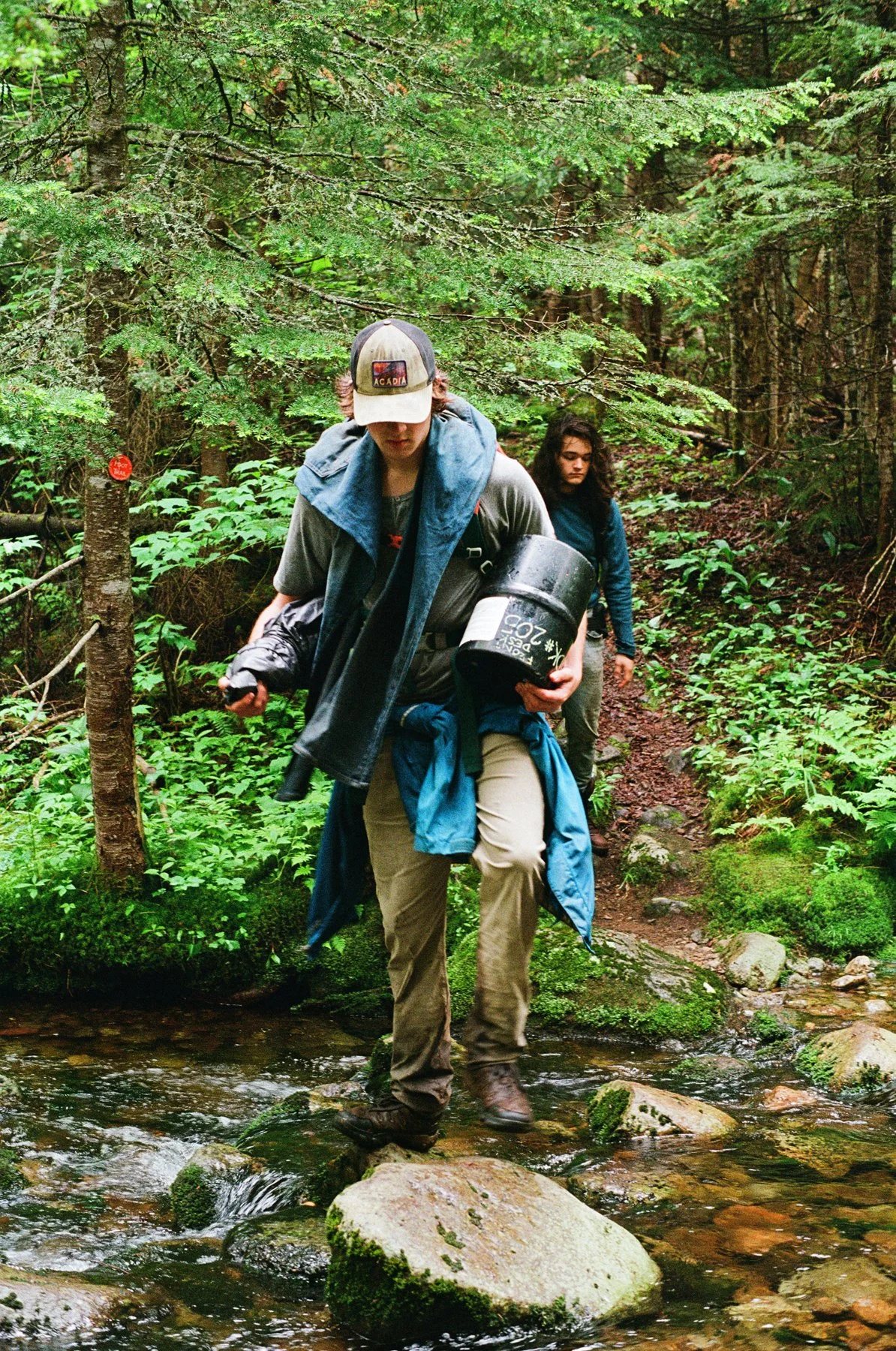 Two people crossing a stream in a forested area, with one person leading and carrying a black container, and another walking behind.