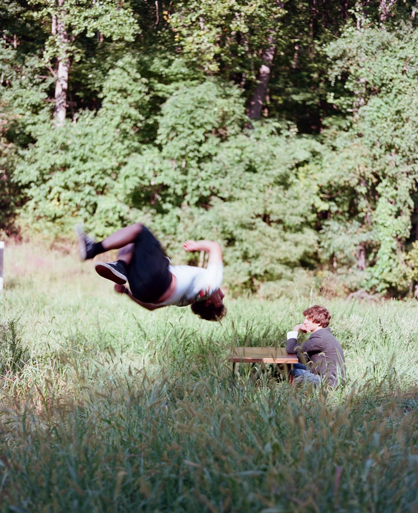 A person doing a backflip in mid-air while another person sits at a wooden table in a grassy field surrounded by trees.