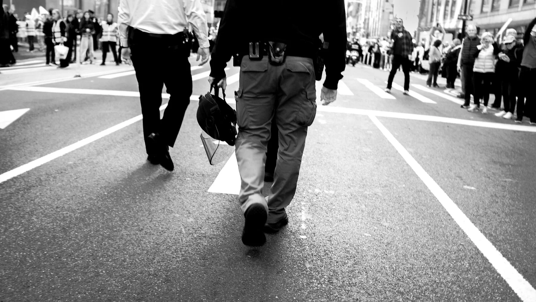 People crossing the street in a busy urban area, with a focus on a person walking away carrying a helmet and a folder.