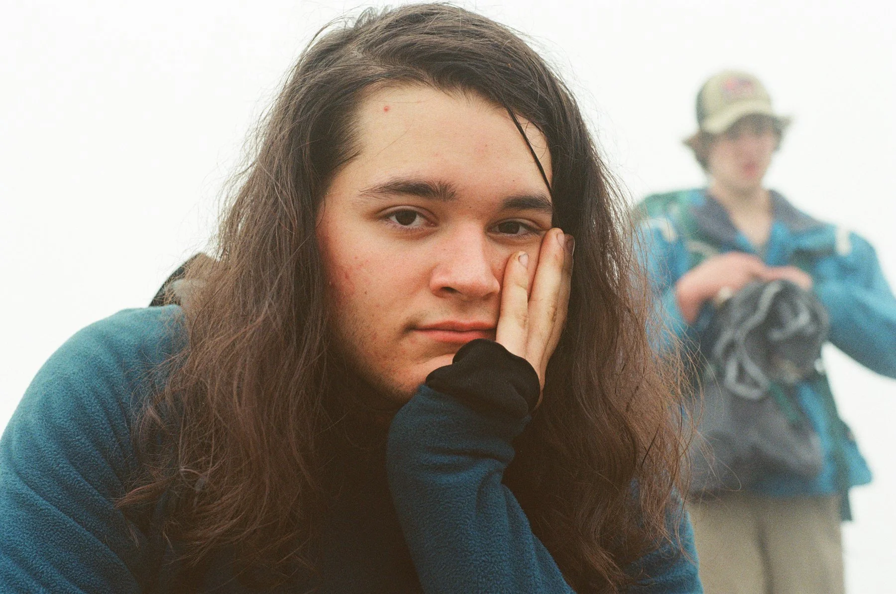 Young person with long dark hair resting their face on their hand, looking into the camera with a serious expression, with another person in the background wearing a blue jacket and hat.