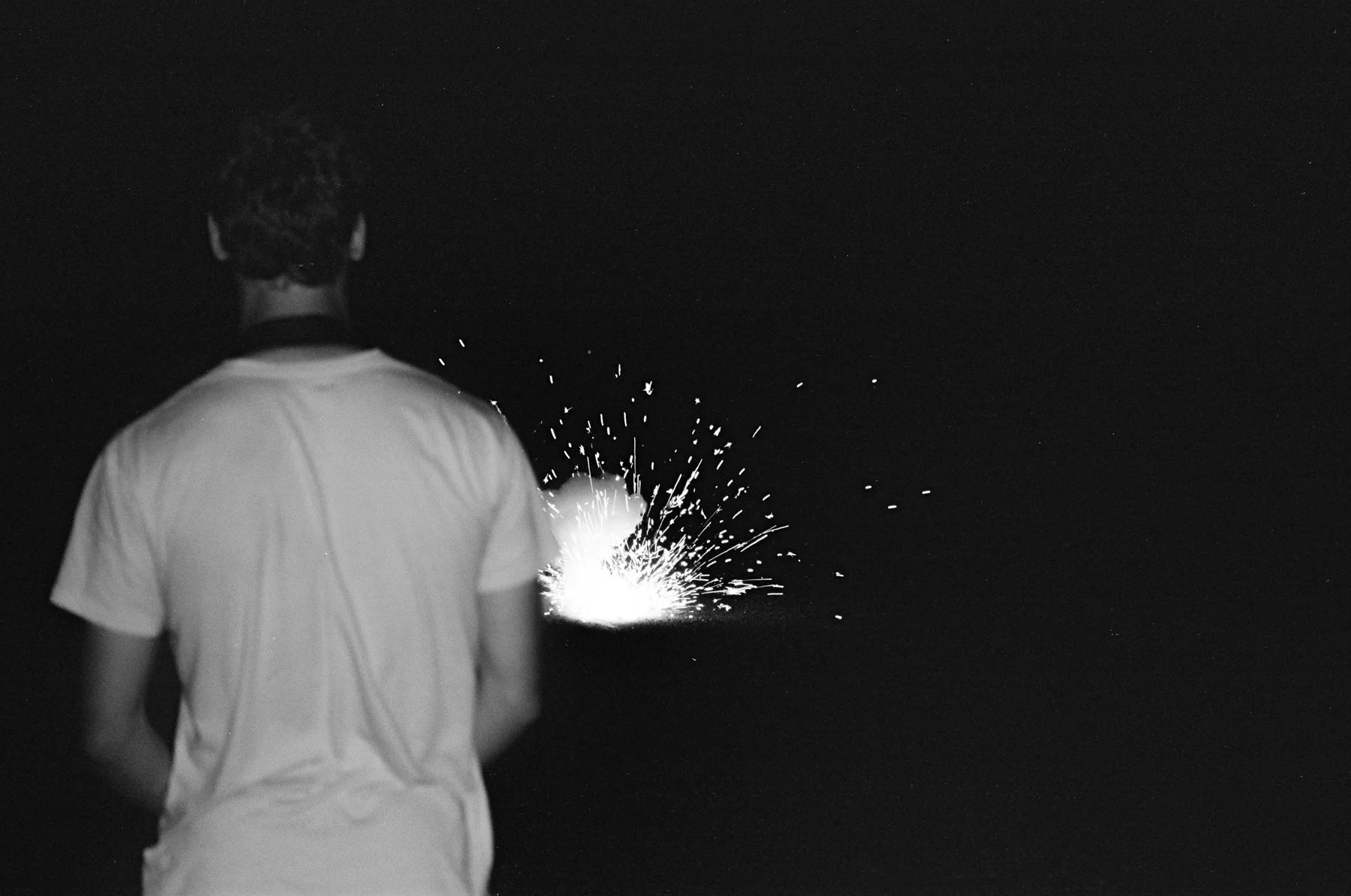 Person standing with their back to the camera, facing sparks flying against a dark background.