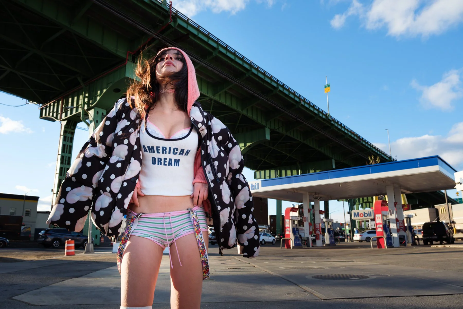 A woman standing at a gas station under a bridge, wearing a polka-dot coat, a white top with 'American Dream' written on it, pink and white striped shorts, and a pink hoodie.
