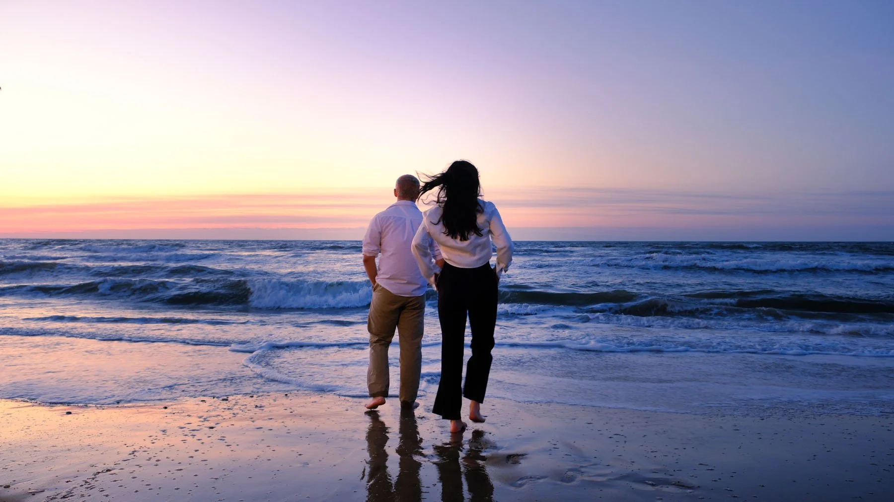 A couple walking along the beach at sunset, with waves crashing behind them.