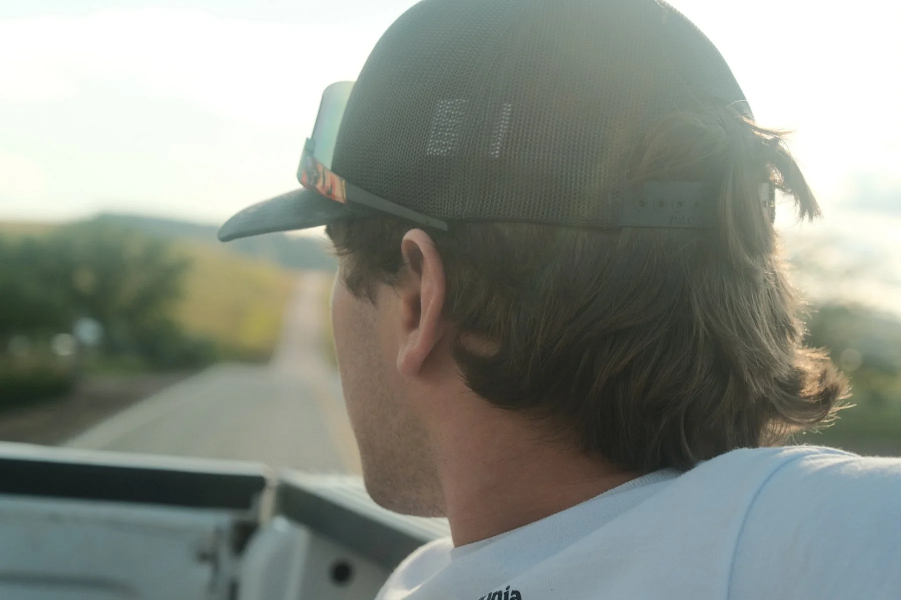 A man with brown hair viewed from behind, wearing a black mesh trucker cap with sunglasses attached, sitting in a vehicle on a rural road with trees and open sky in the background.