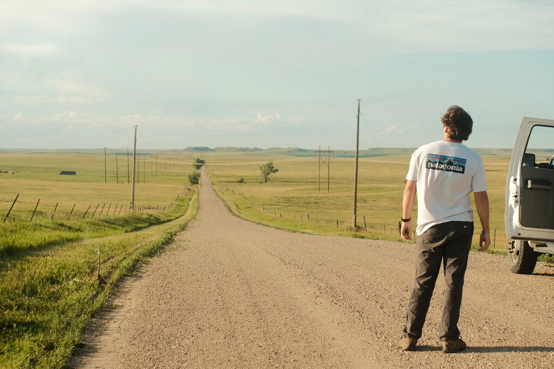 A person walking along a gravel country road with open grassy fields on either side, under a partly cloudy sky, wearing a white Patagonia t-shirt and dark pants.