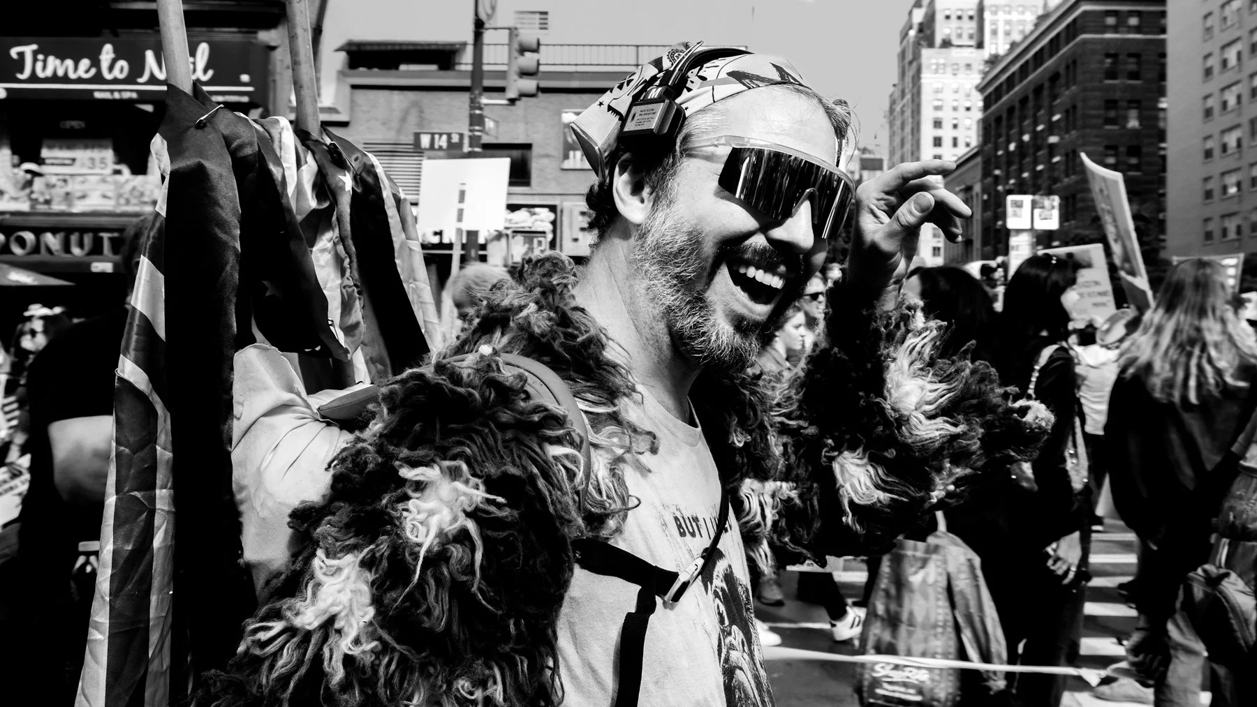 A man with a beard and curly hair wearing sunglasses, a headband, and a furry jacket, smiling and holding an American flag while participating in a crowd protest or demonstration in an urban area.