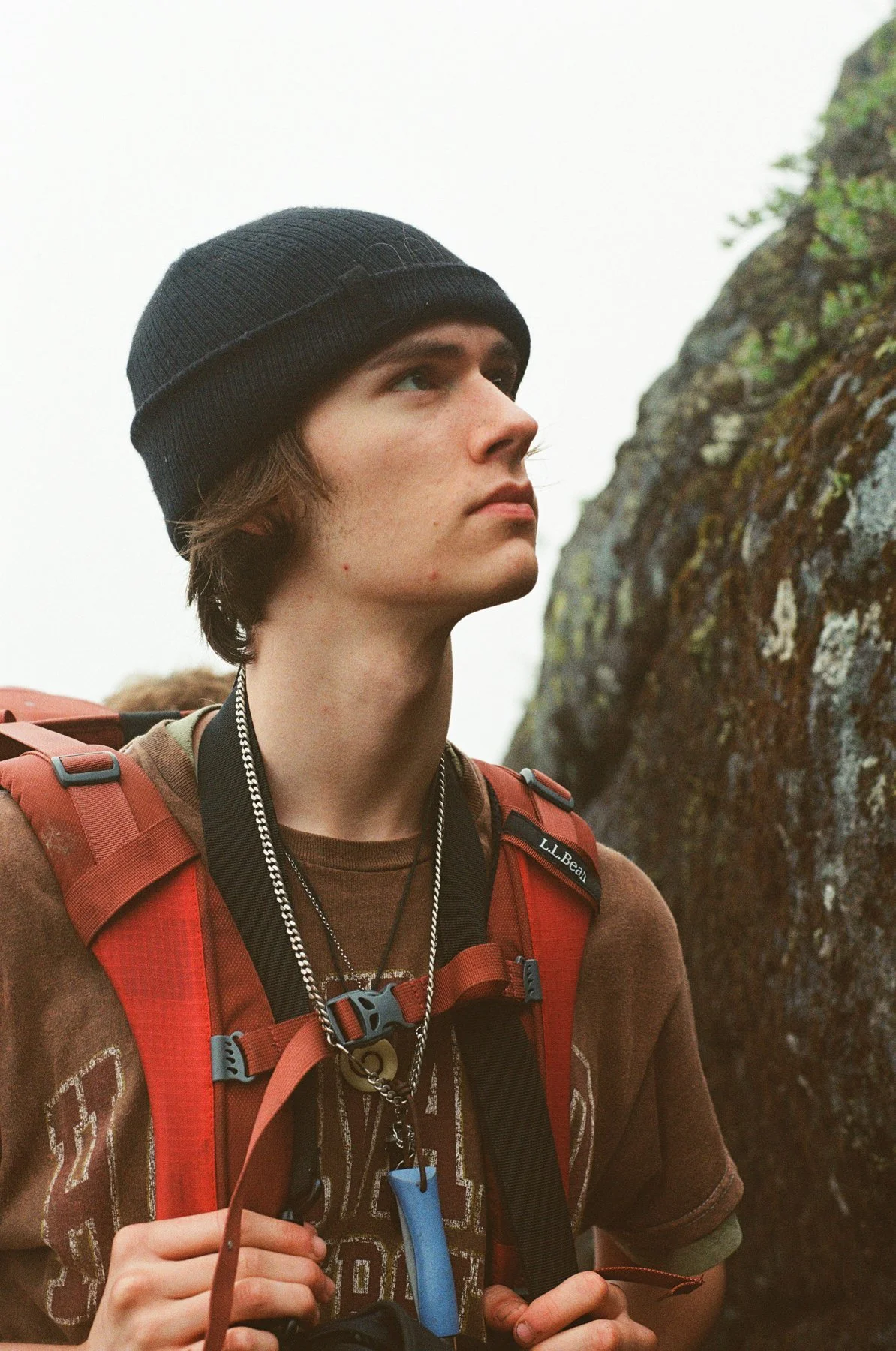 A young man with brown hair, wearing a black knit beanie and a brown shirt, carrying a red hiking backpack with various accessories, stands outdoors near a moss-covered rock formation, looking off into the distance.