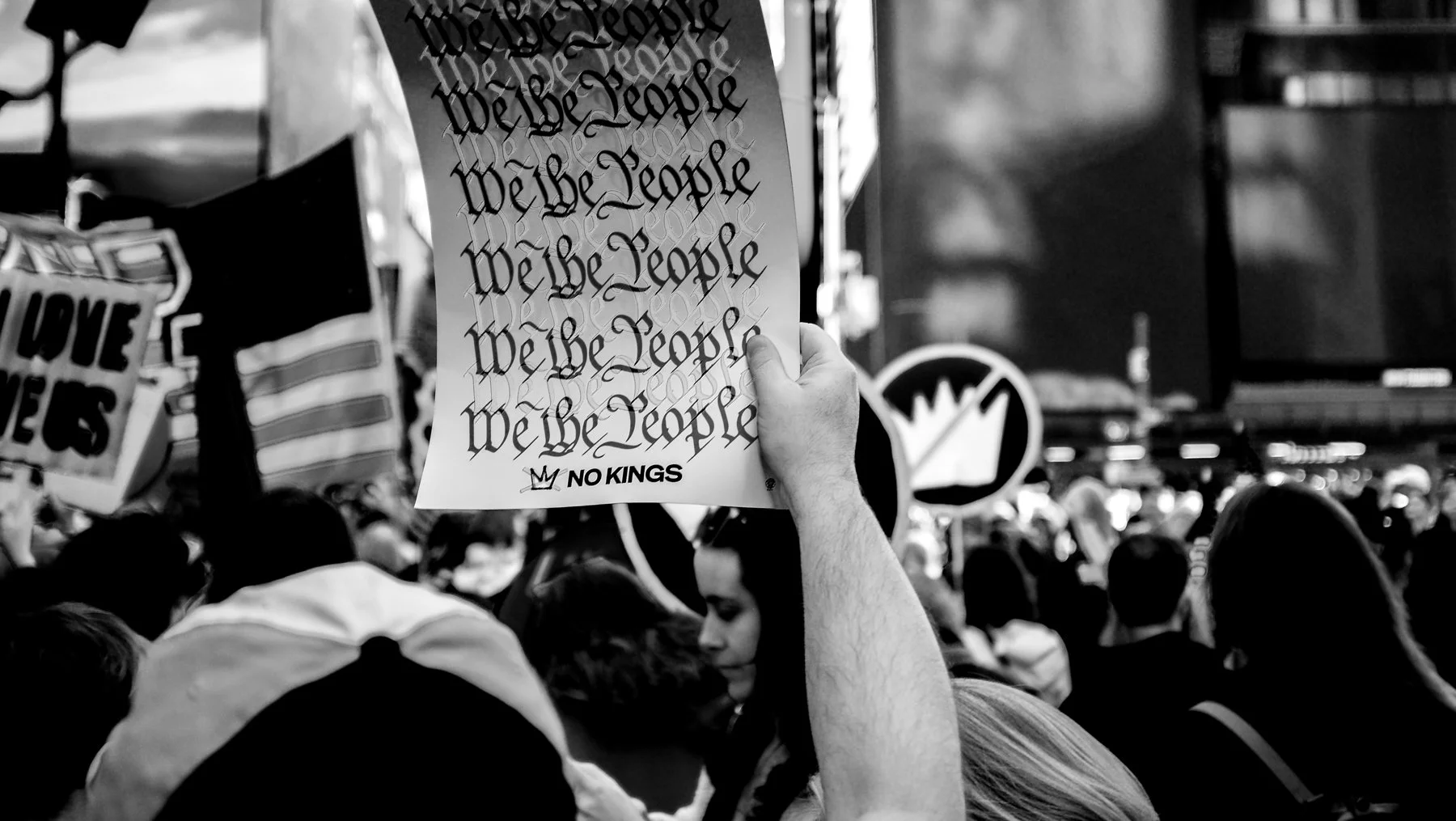 A crowd of people at a protest or demonstration, with a hand holding a sign that reads "We the People" in stylized script and "NO KINGS" at the bottom, in black and white photography.