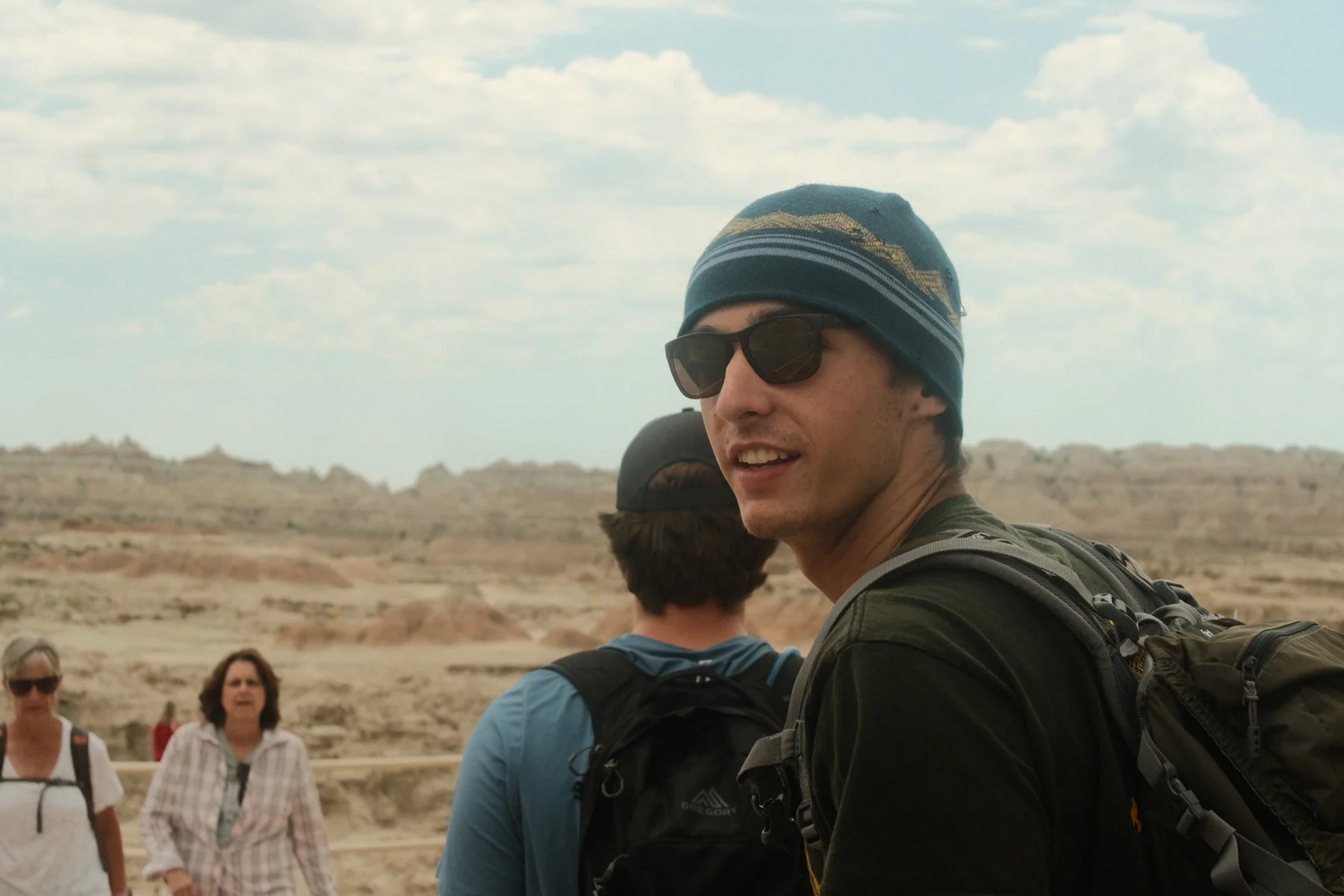 A man wearing sunglasses, a beanie, and a backpack, standing outdoors with desert landscape and other tourists in the background.