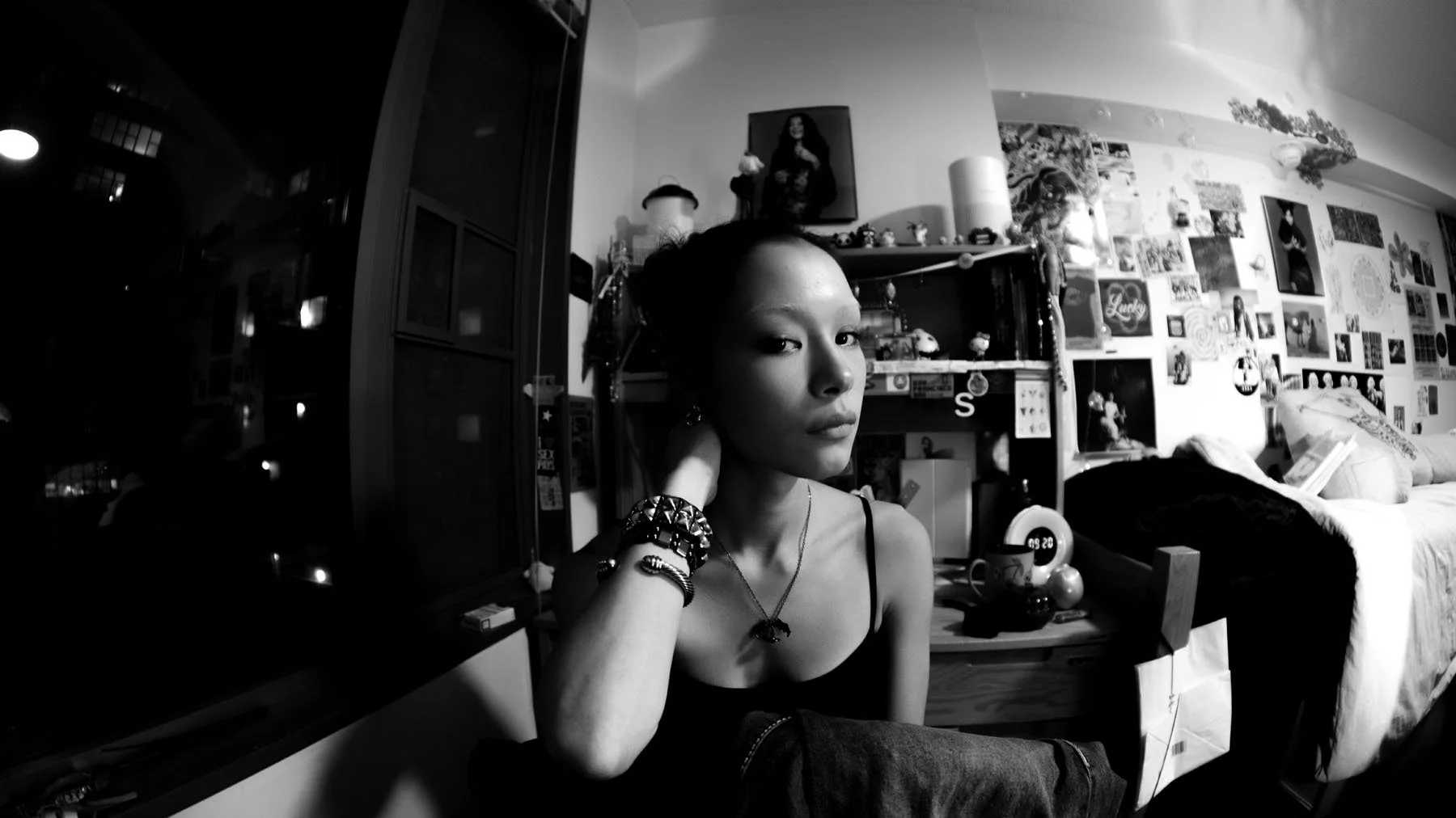 A young woman with braided hair, wearing a tank top and jewelry, sitting in a decorated room with posters and photos on the wall, a bed, and various personal items.