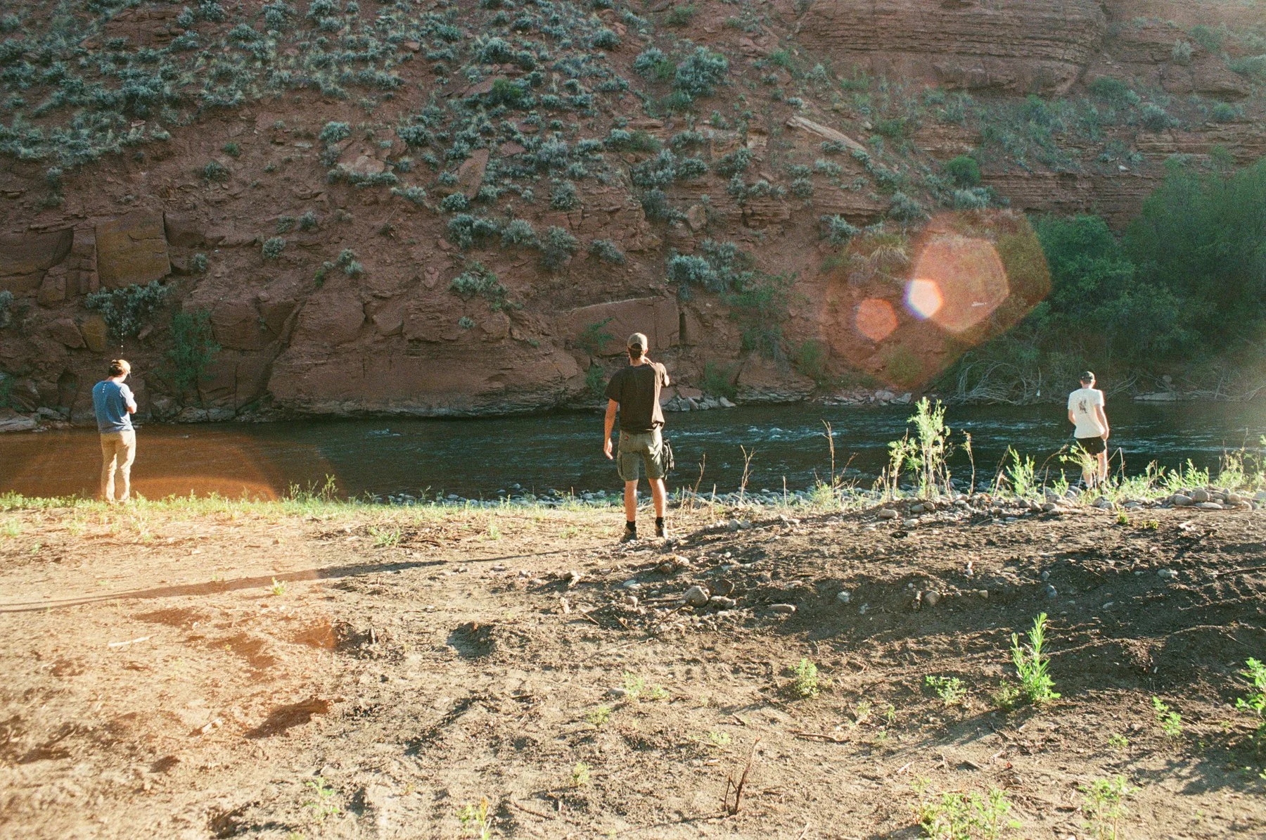 Three young men standing on the sandy riverbank, looking out over a river with a rocky and plant-covered canyon in the background.