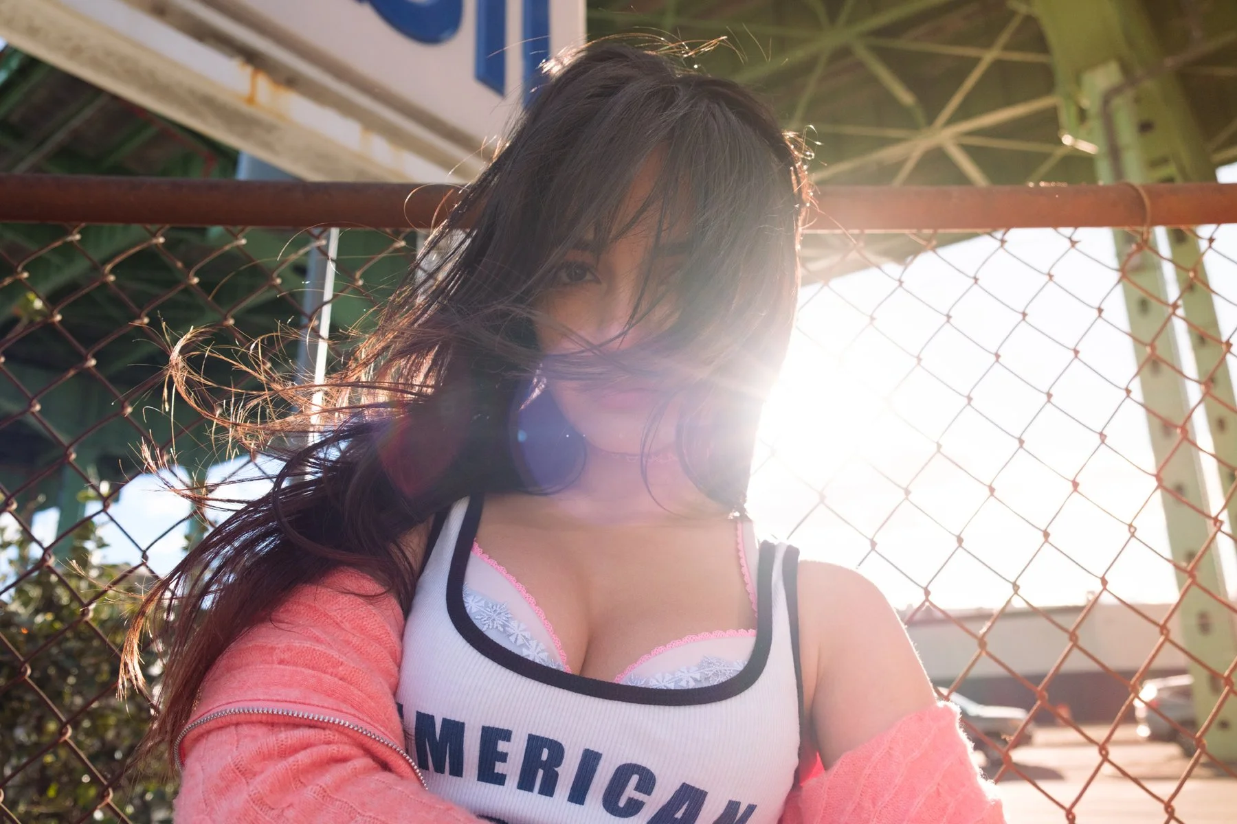 Young woman with long dark hair standing near a chain-link fence, sunlight shining behind her, wearing a white top with 'AMERICAN' text and pink accents, pink sweater off her shoulders.