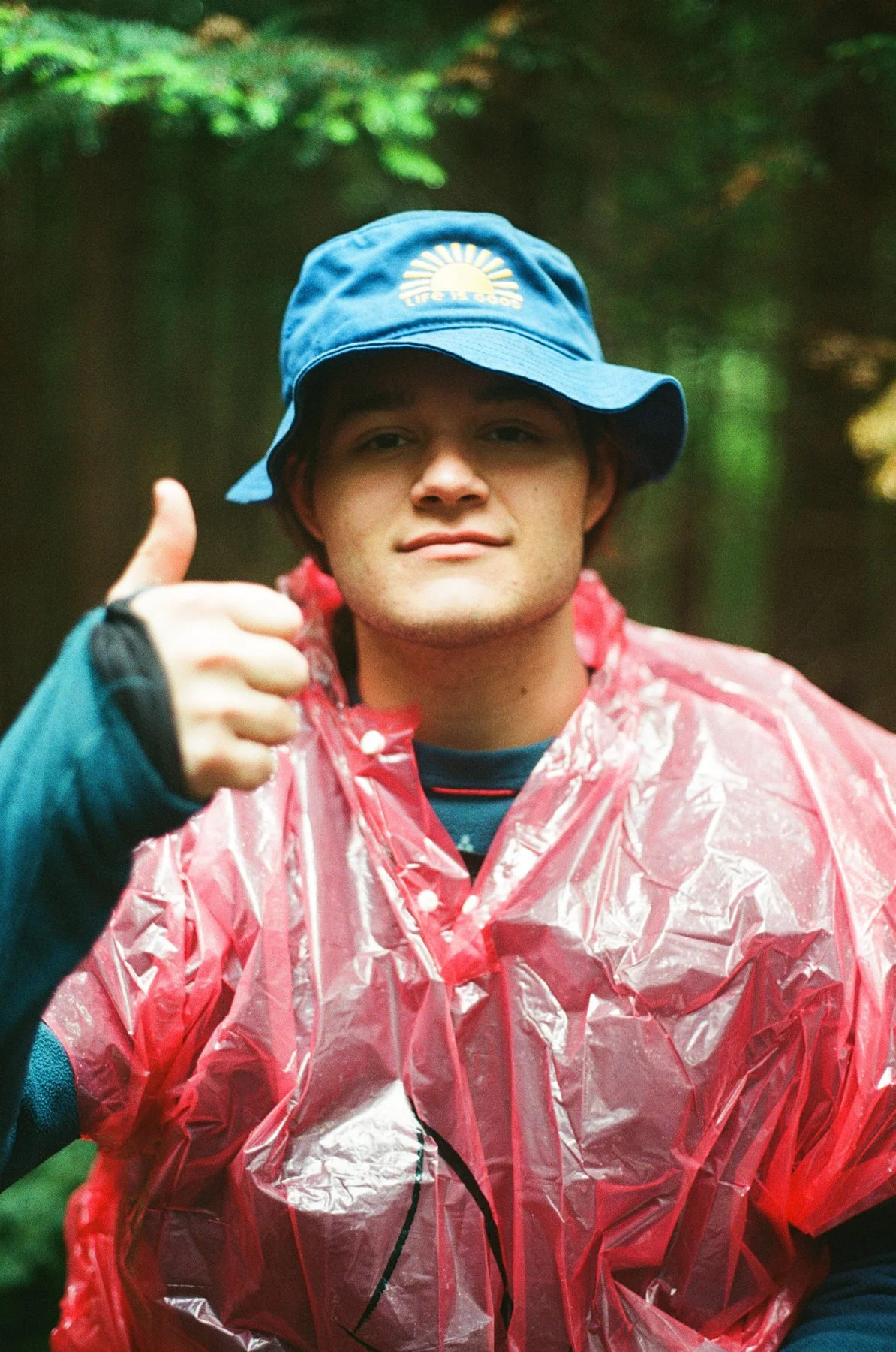 A young man wearing a blue bucket hat with a "Life is Good" logo, a pink rain poncho, and giving a thumbs-up in a forested area.
