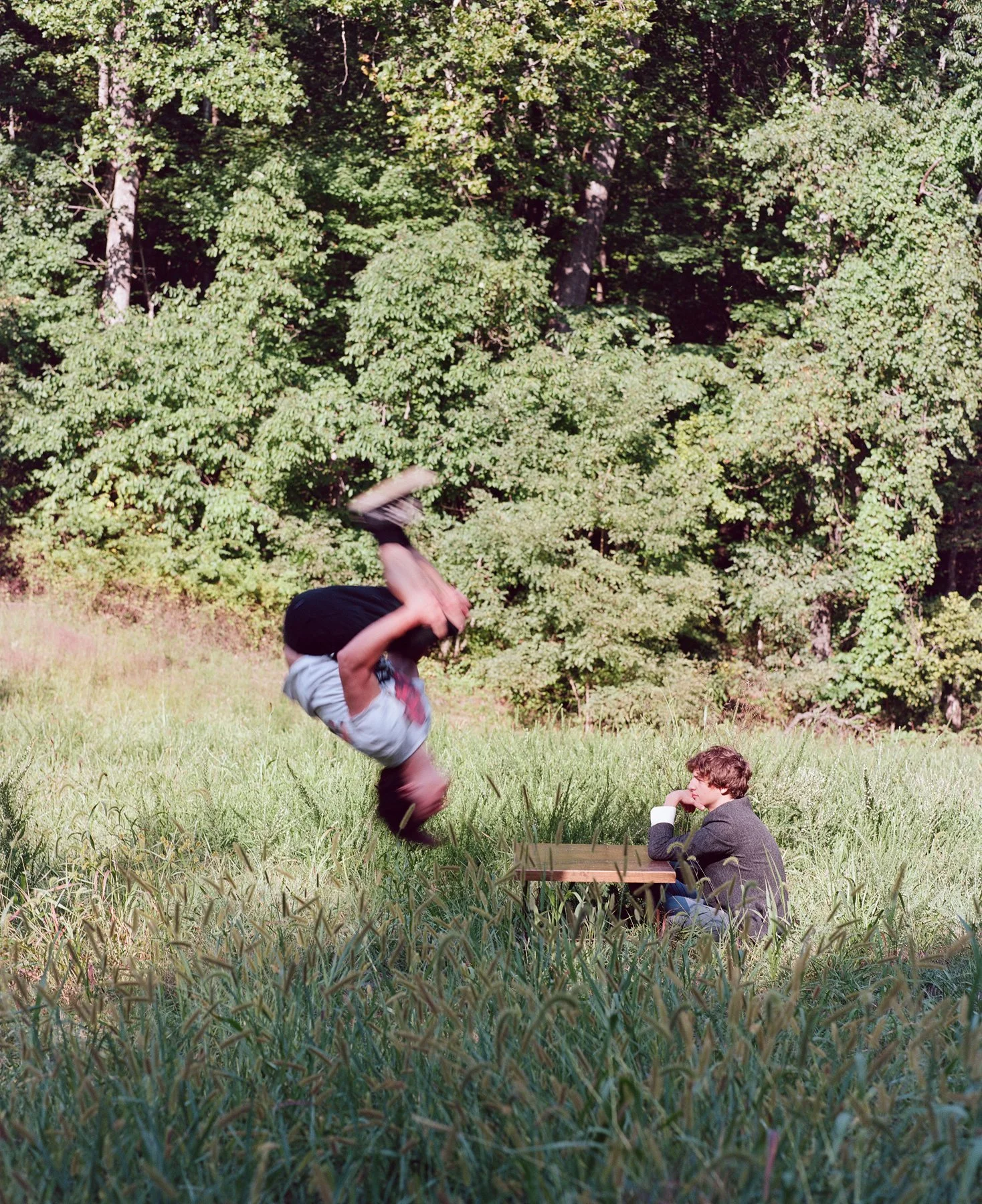 A person in mid-air flip over a seated man at a picnic table in a grassy field with trees in the background.
