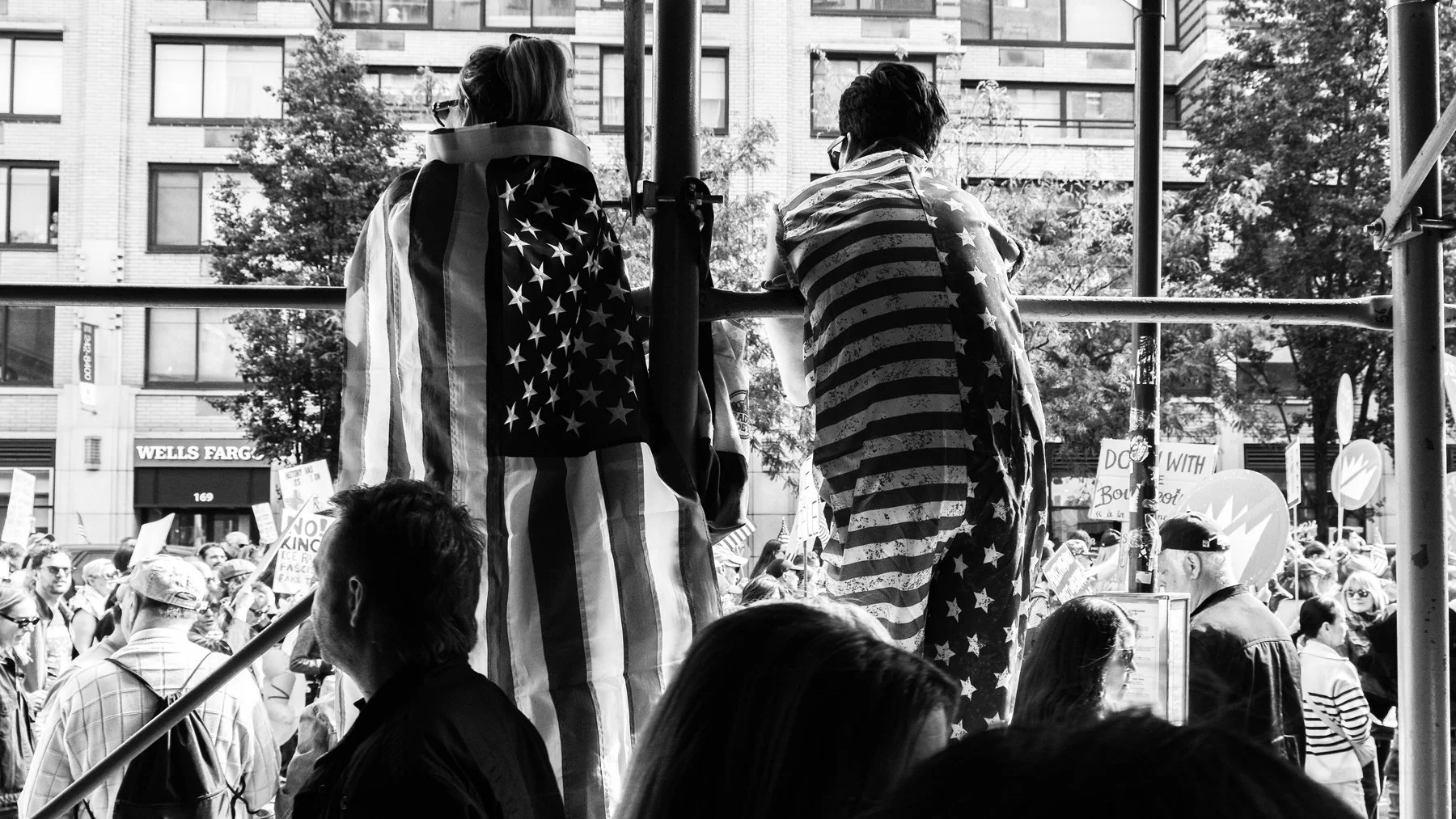 Two individuals standing on a platform at a protest, covered in American flag blankets, with a large crowd of protesters in the background holding signs and flags.