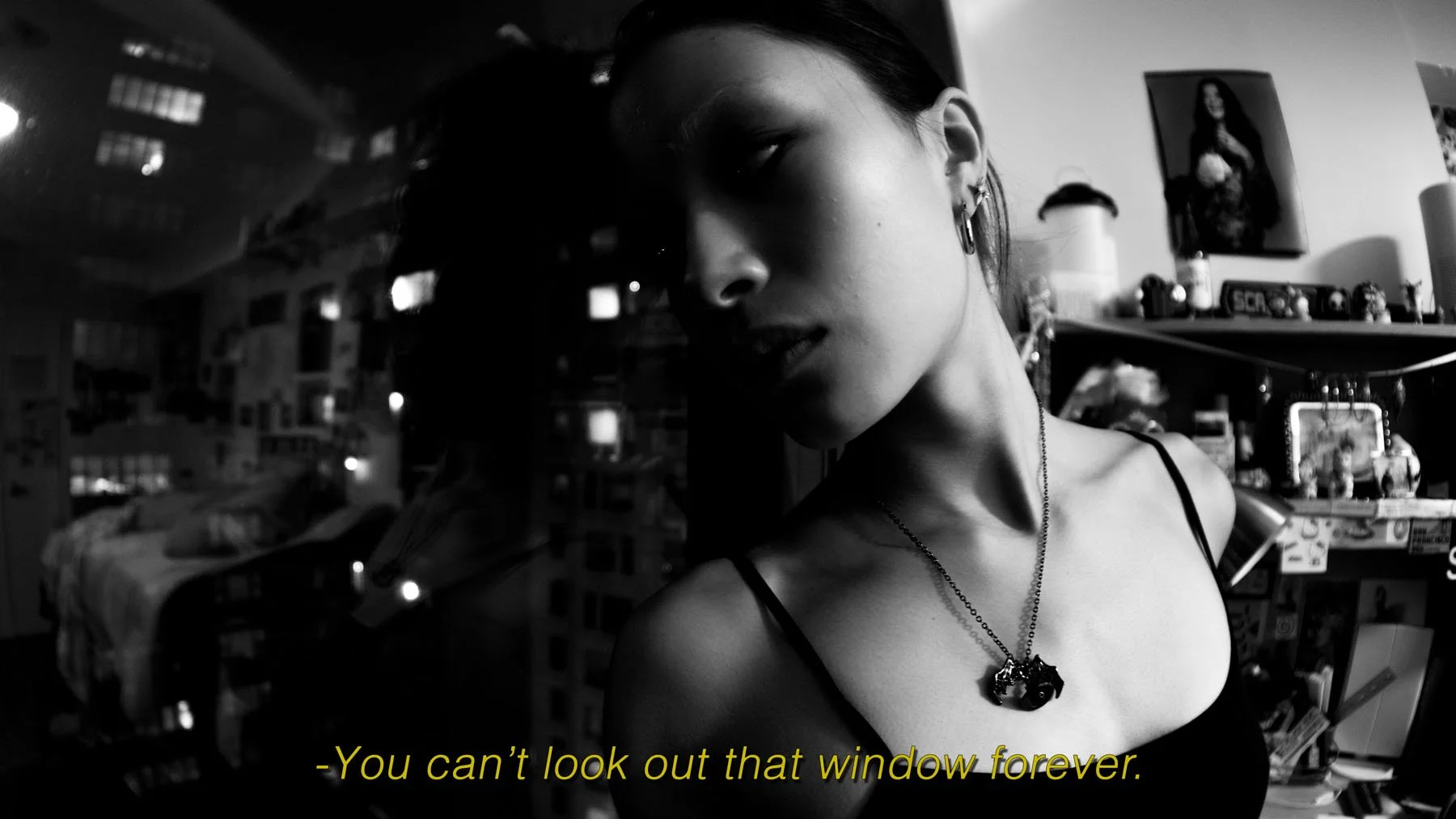 A black and white photo of a woman with dark hair wearing a tank top, earrings, and a necklace, looking over her shoulder inside a room with shelves and pictures on the wall, with the caption 'You can't look out that window forever.'