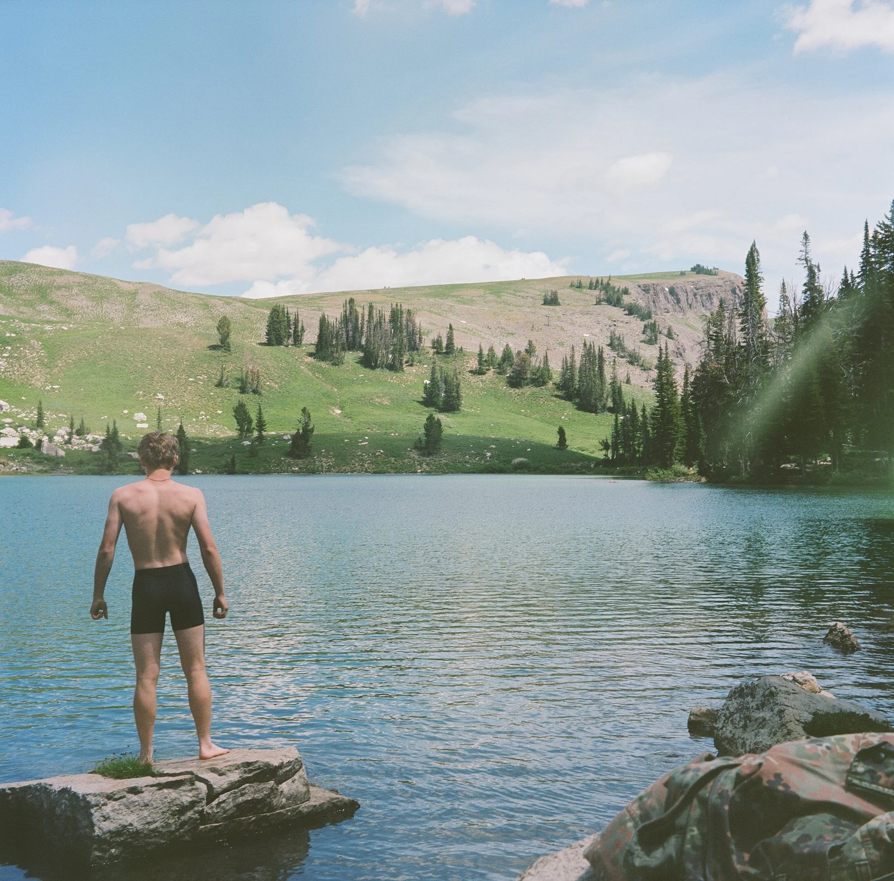Young man standing on a rock at the edge of a lake, looking at the water with hills and trees in the background on a bright, partly cloudy day.