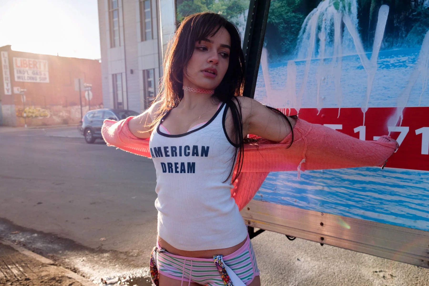 Young woman with long dark hair stretching her arms in front of a colorful sign on a city street, wearing a white tank top with blue text, pink and white striped shorts, and a pink mesh long-sleeve shirt.