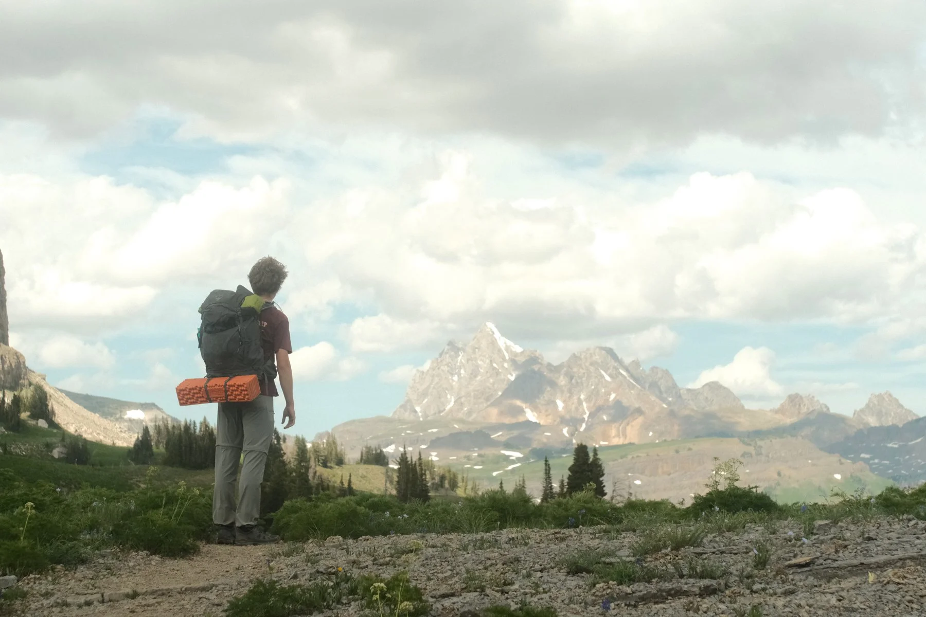 A hiker with a backpack and a foam sleeping pad attached, standing on a trail and looking at a mountain range in the distance under a partly cloudy sky.