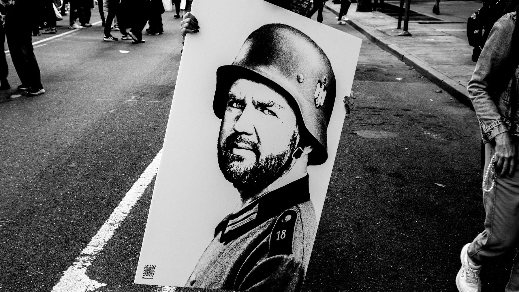 A person holding a protest sign featuring a black-and-white image of a man in a military uniform wearing a helmet, with a serious expression. The protest is taking place on a street with several other people visible in the background.