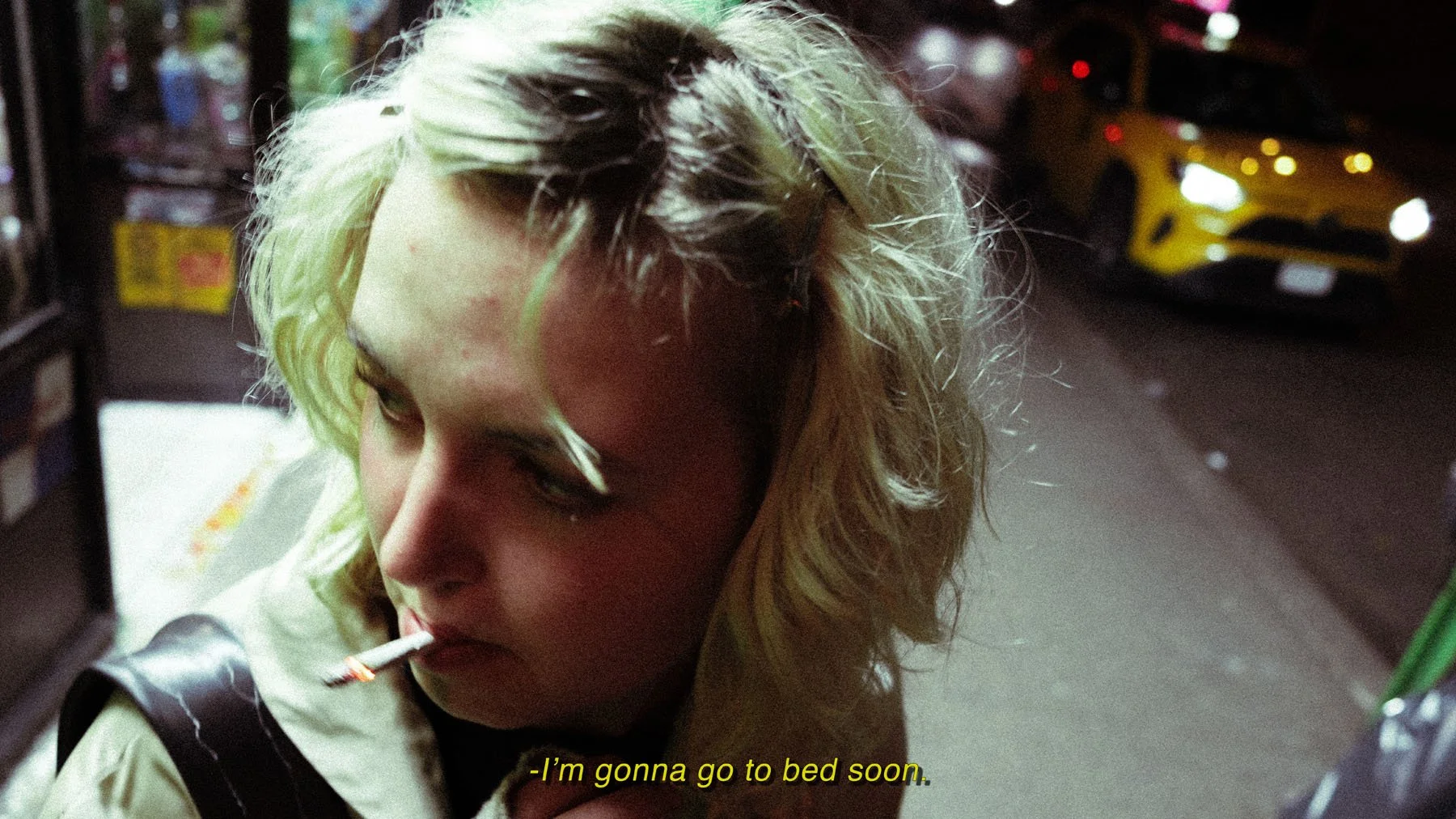 Close-up of a woman with blonde, messy hair smoking a cigarette at night on a city street, with a yellow taxi and storefronts in the background. Subtitles read, "I'm gonna go to bed soon."