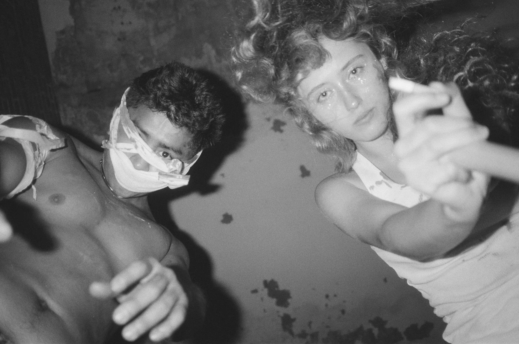 Black-and-white photo of a man wearing a mask and goggles, shirtless, and a woman with curly hair, holding a camera, both looking at the camera.