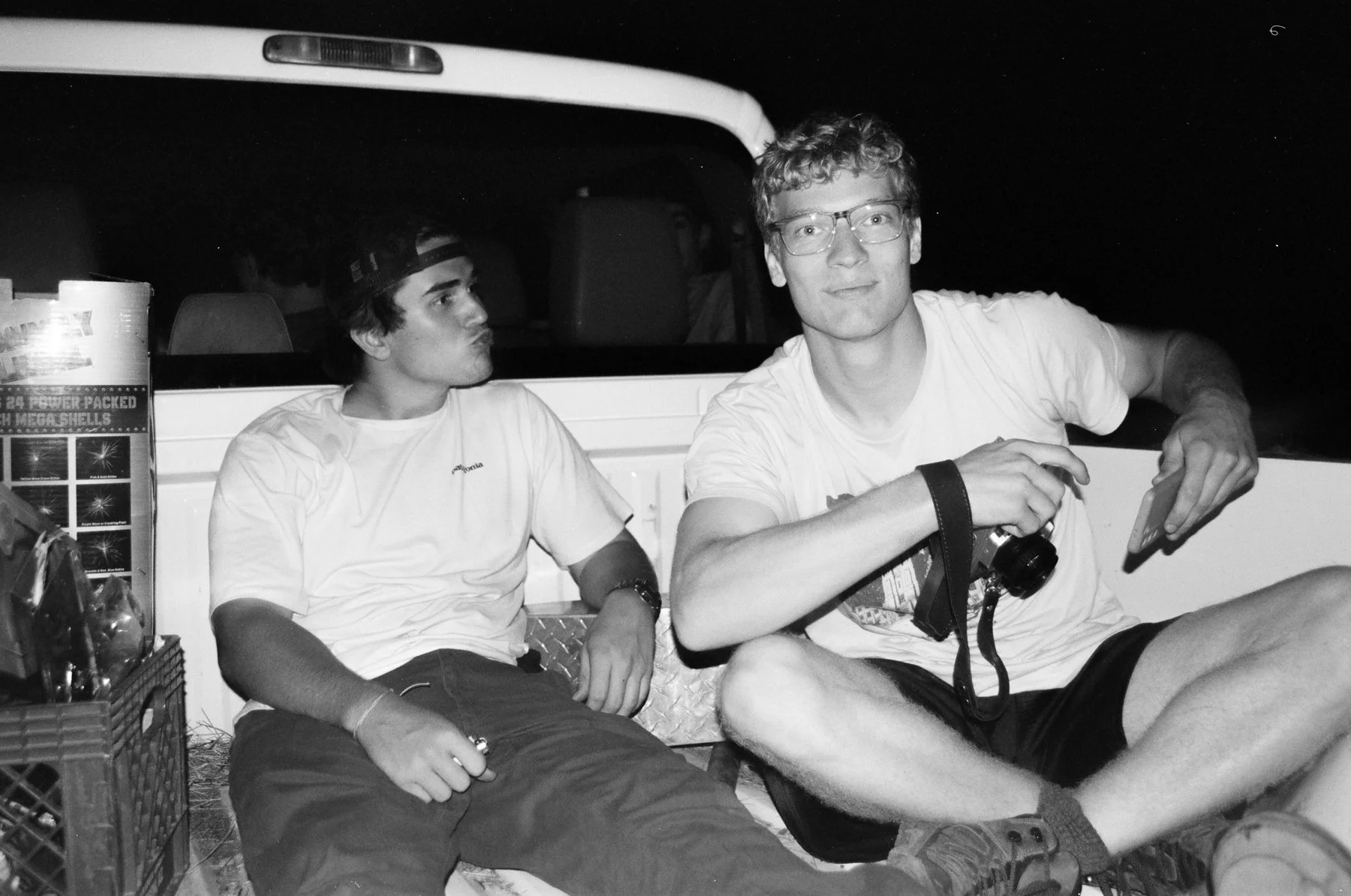 Two young men sitting on the back of a pickup truck at night, one is holding a camera and a phone, and the other has a cigarette in hand, with fireworks on a box beside them.