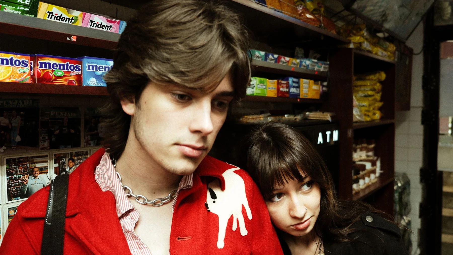 A young man and woman in a grocery store aisle, with the man wearing a red jacket with a white dog patch on the shoulder and a chain necklace, looking down, and the woman leaning on his shoulder with her face partly visible, looking to the side.