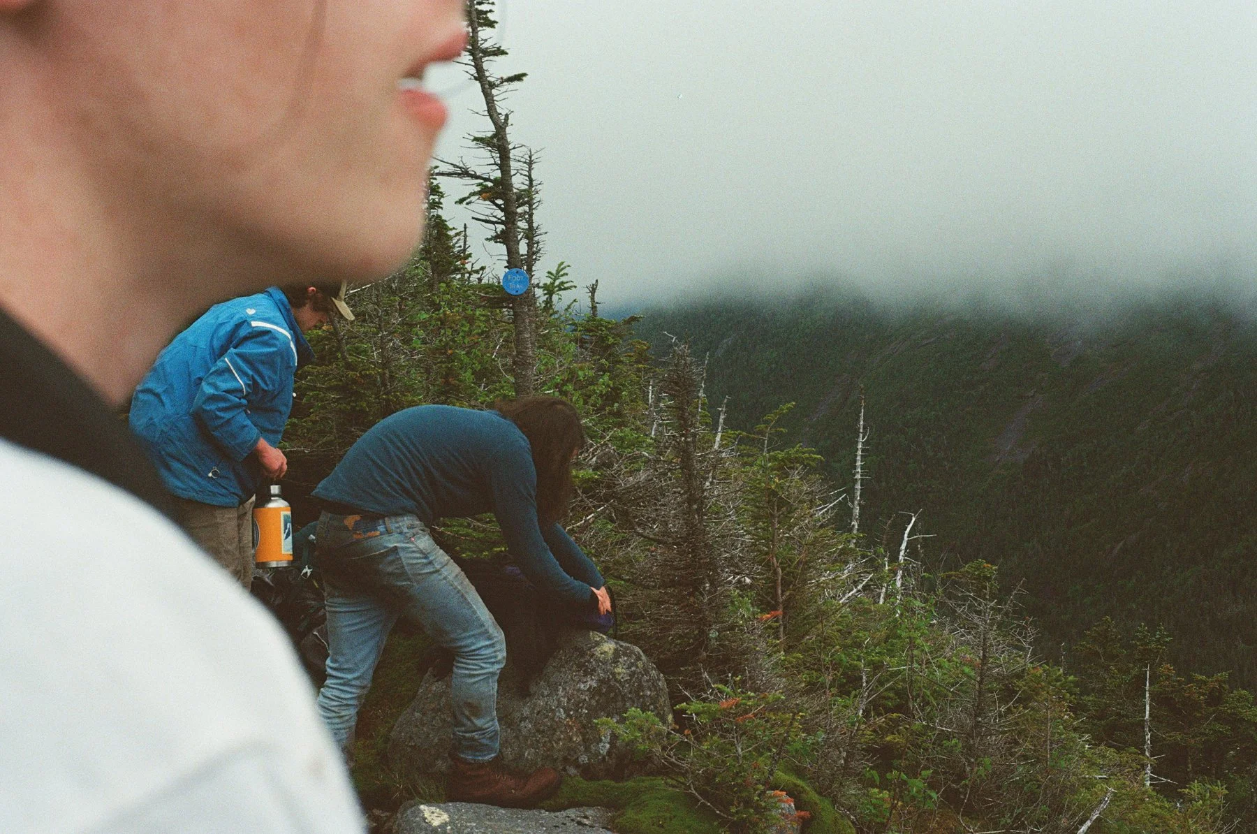 Close-up of a person's face in the foreground with three people on a mountain trail near a forested area in the background.