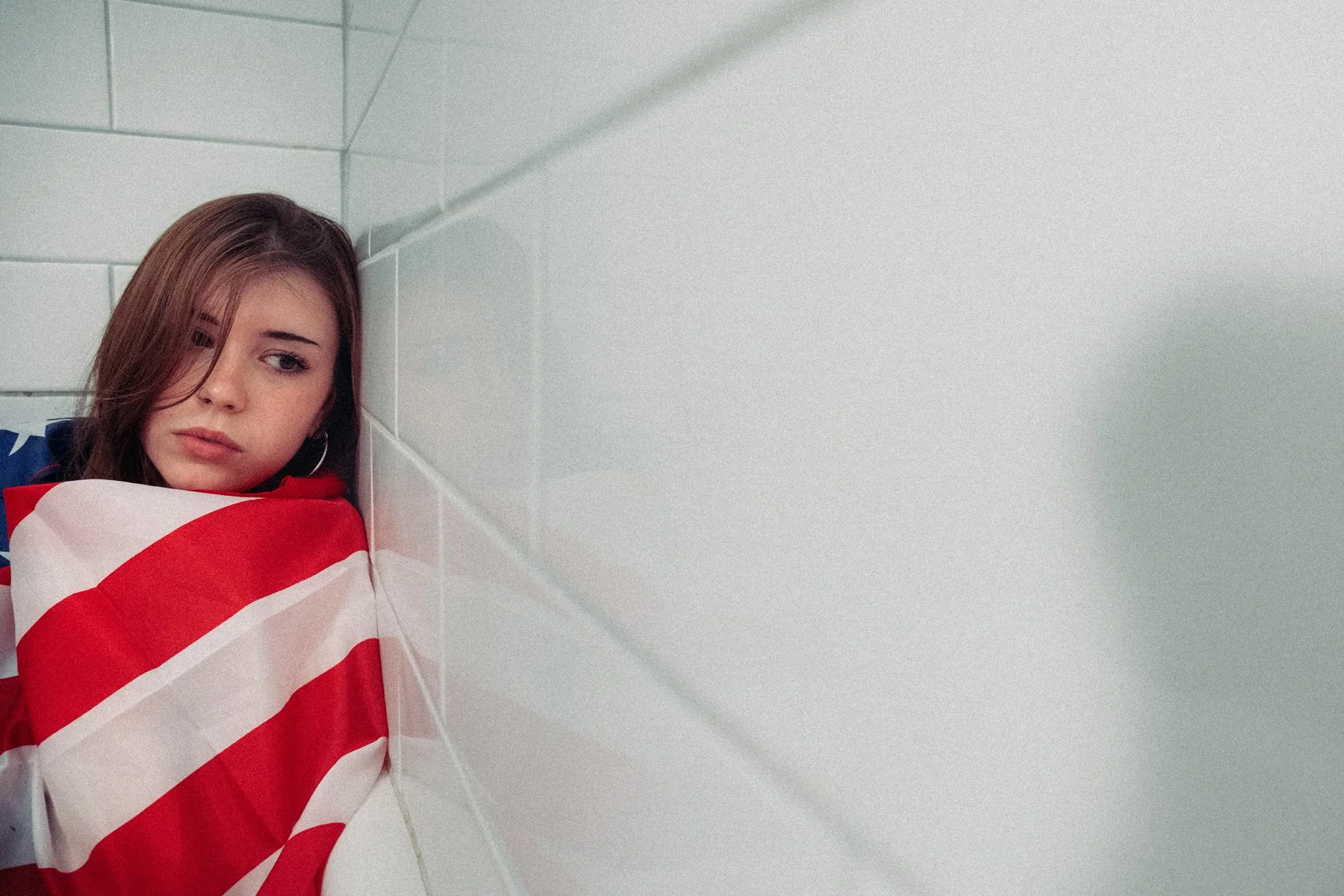 Young woman with brown hair and hoop earrings in a bathroom, wrapped in a red and white striped flag, leaning against tiled wall, with a somber expression.