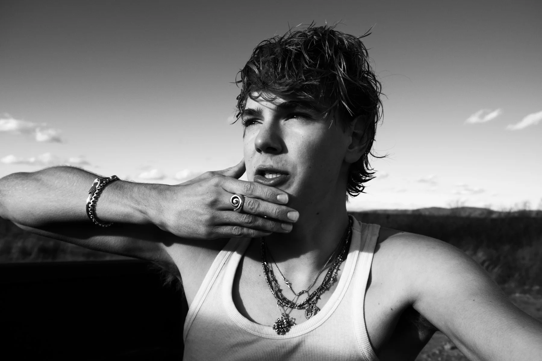 Black and white photo of a young person with tousled hair, wearing layered necklaces and a tank top, holding one hand to their face, outdoors with a landscape and sky in the background.